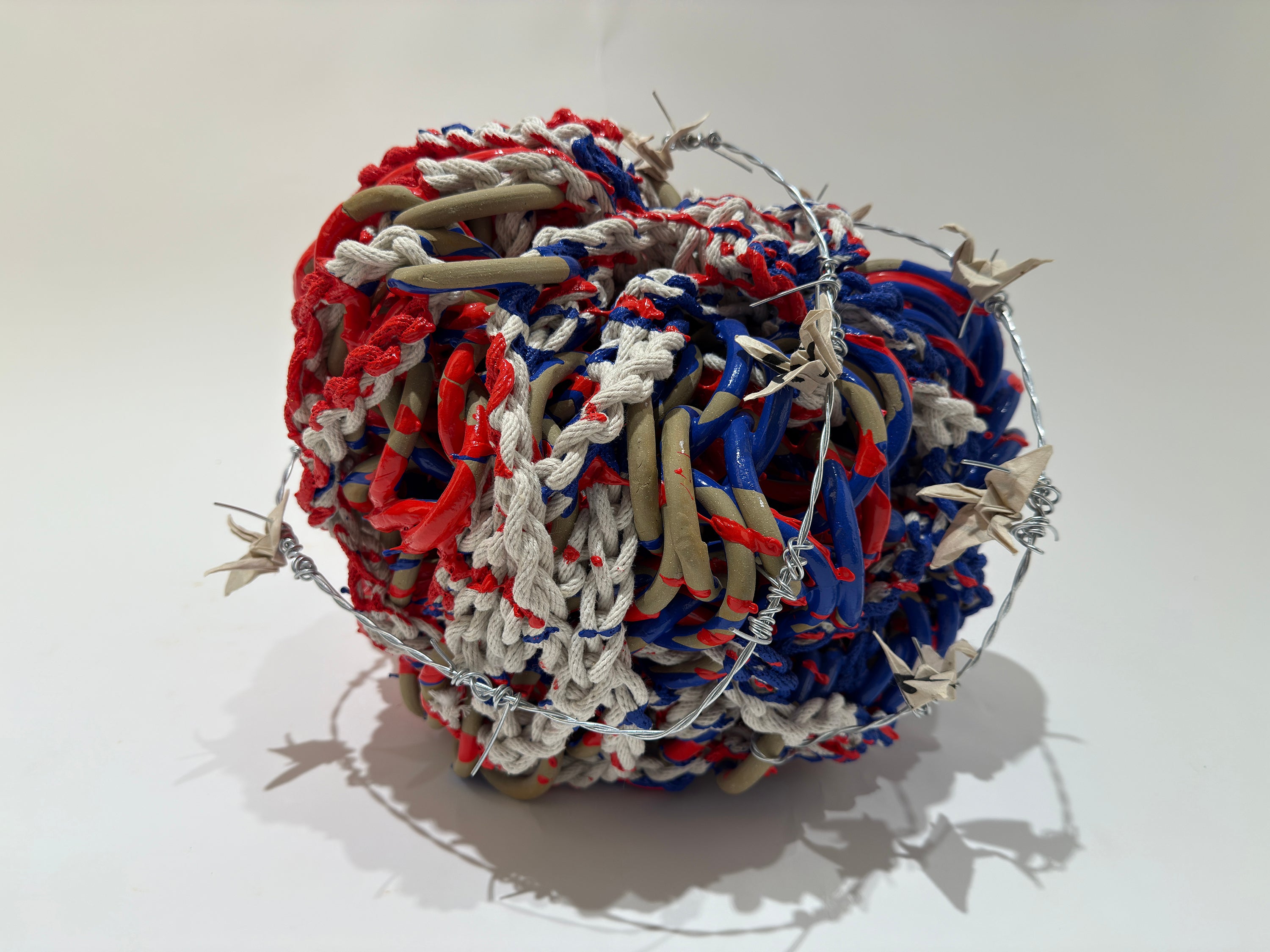 Loops smooth tan chainmail are constricted by white rope.  The piece is bathed in red and blue paint and paper cranes are spears by wrappings of barbed wire around the piece.