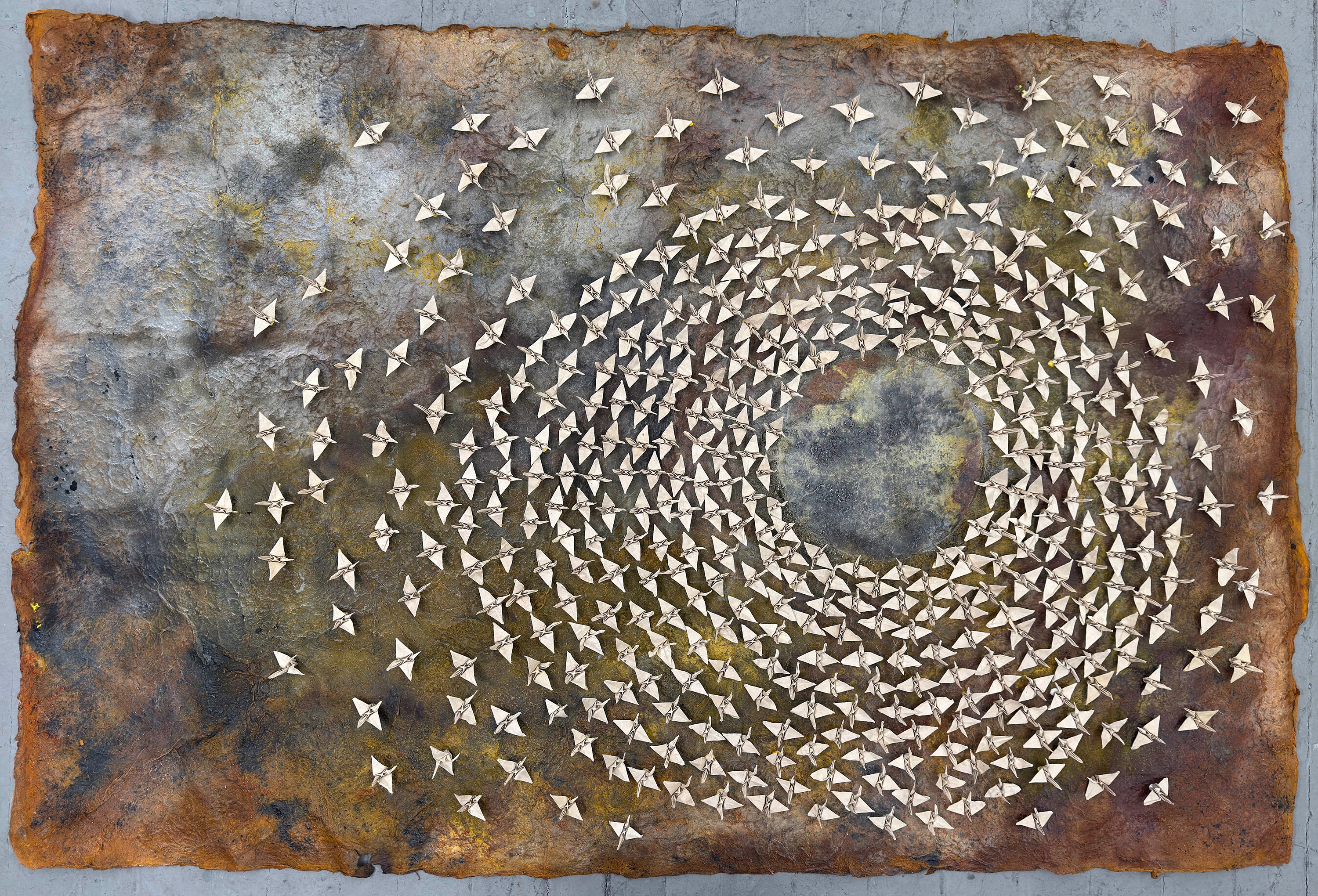 A swarm of paper cranes fixate on a circle within the large piece of paper.  The cranes are pure and white while the paper they sit upon is worn, dyed with age, and at rick of colapse.