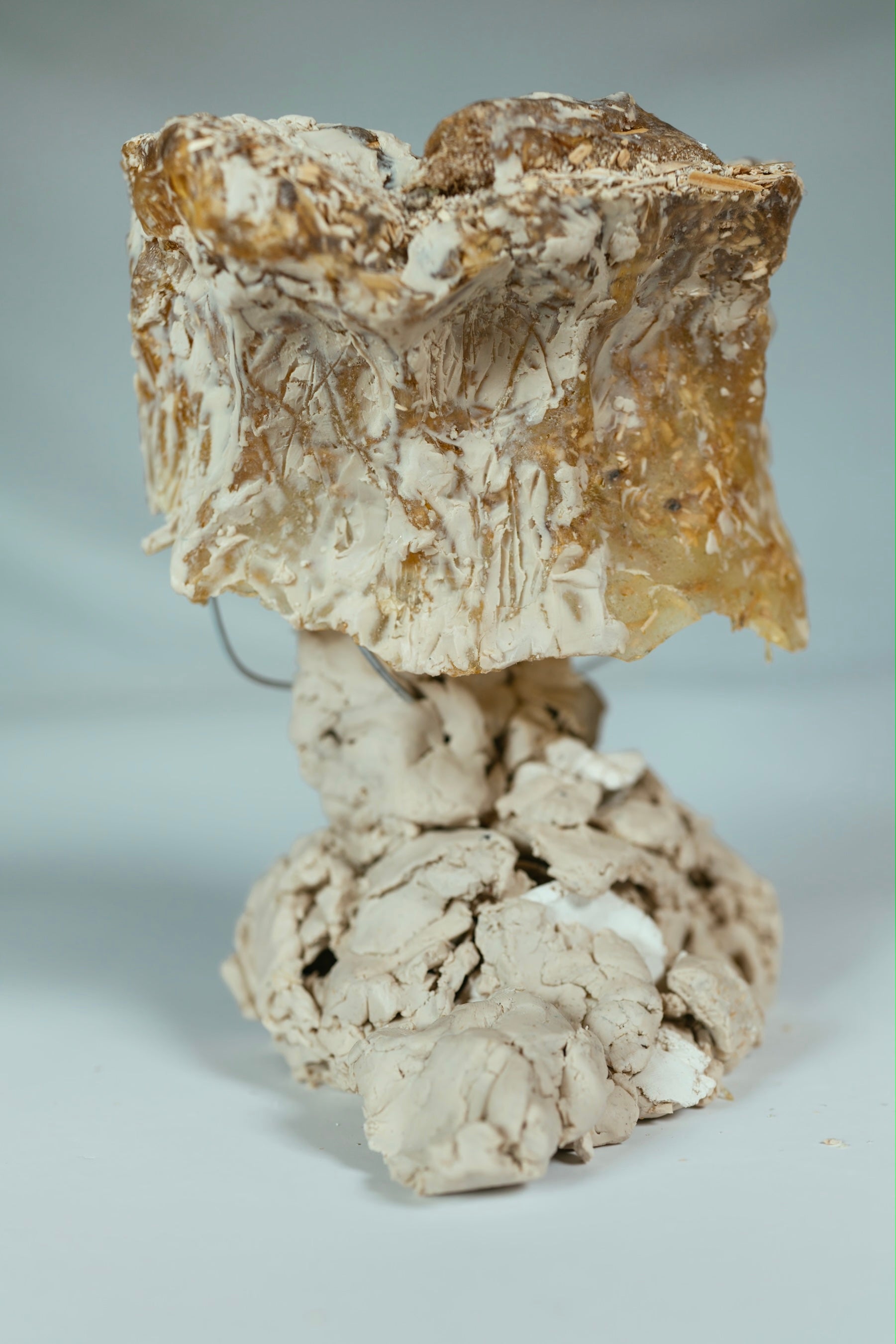 An experimental hemp and pine-rosin lamp shade resting on a hand-molded clay base. The amber translucent biocomposite shade shows organic folds and textures from the clay-forming process.