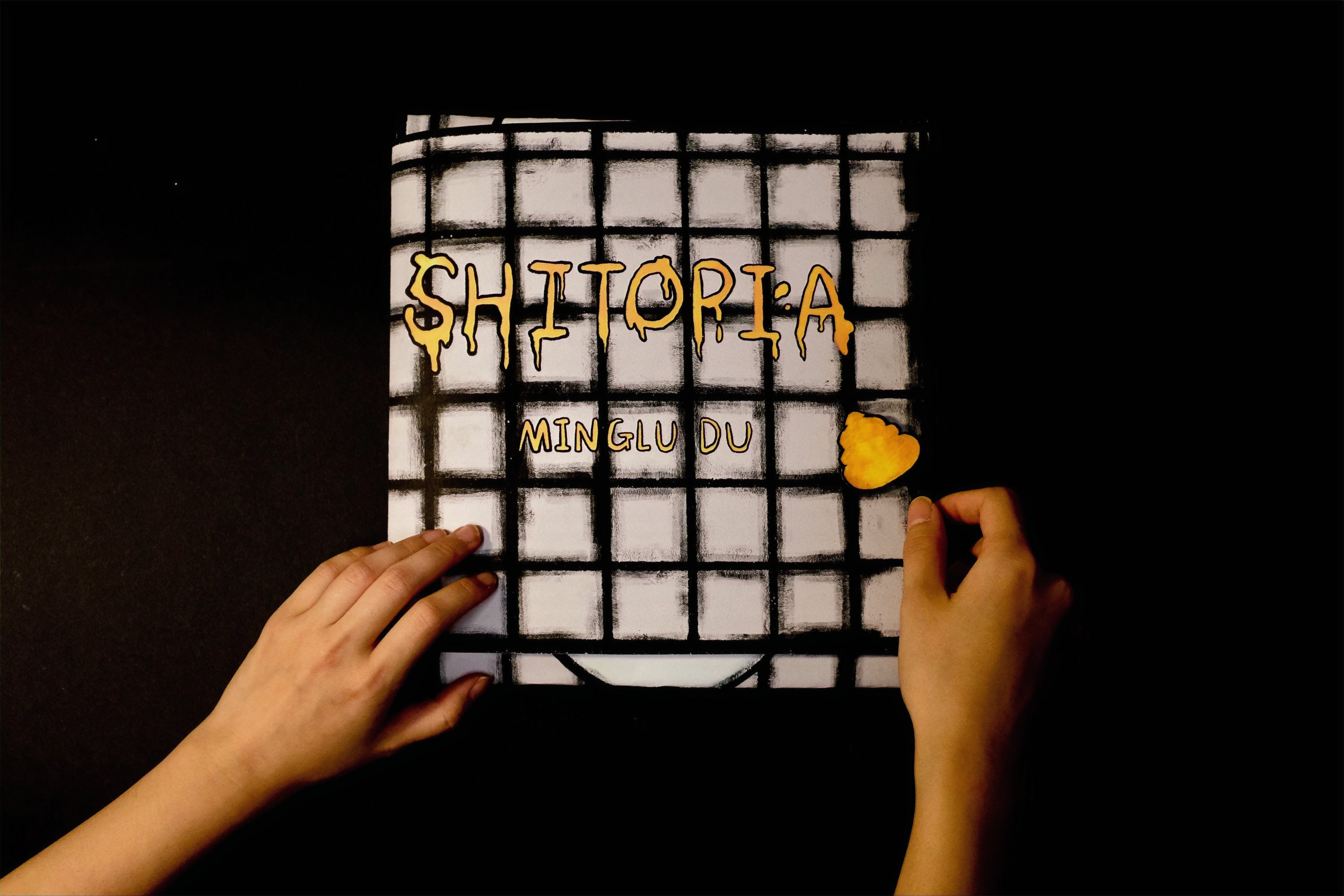 The cover of the comic book Shitopia is designed as a foldable outer shell shaped like a tiled toilet and toilet seat that wraps around the book’s contents. The cover includes the title of the work, the author’s name, and a pop-up paper clasp shaped like a piece of excrement that can be tied with string to keep the book closed. The handmade structure combines absurd humor and tactile sculptural elements.
