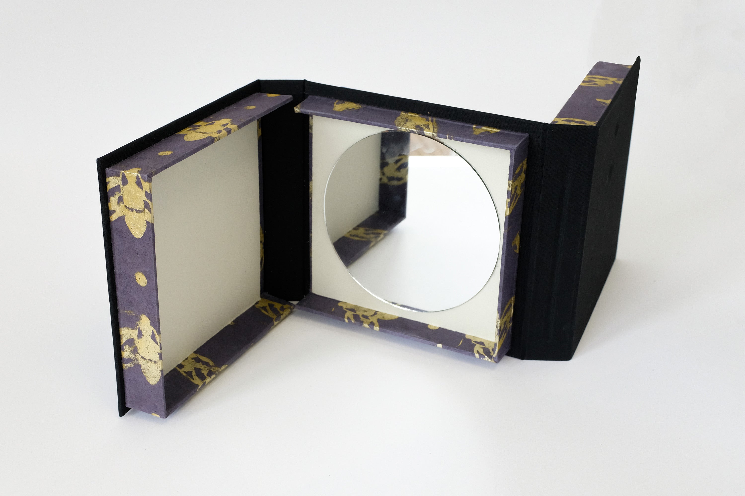 Double sided box against a white background that has a mirror inside of it. The edge of the box has a gold moth pattern.