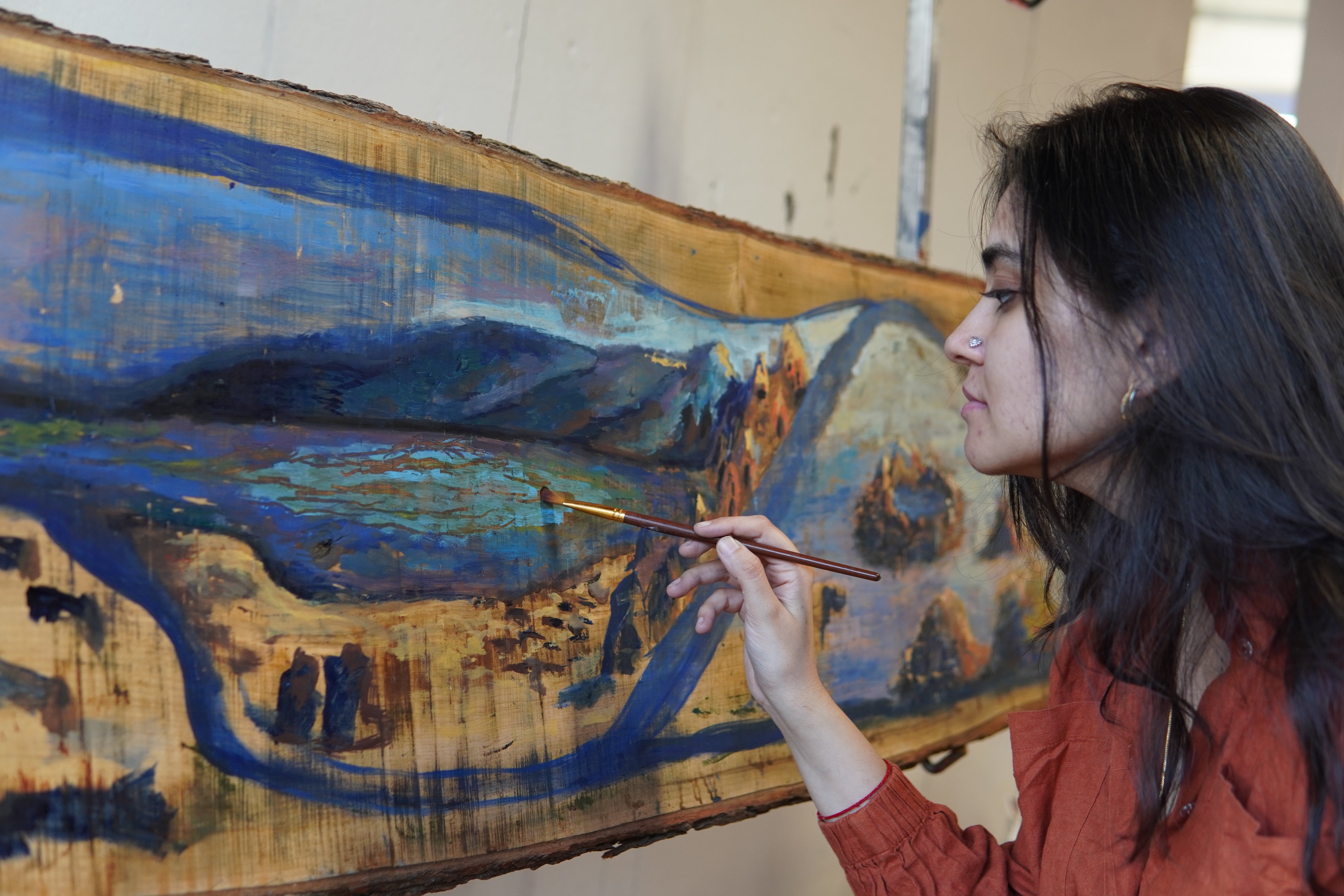 A South Asian woman with long dark hair and a nose ring holds a fine paintbrush close to a large live-edge maple panel propped at an angle. The panel is painted in deep blues, warm ochres, and muted greens