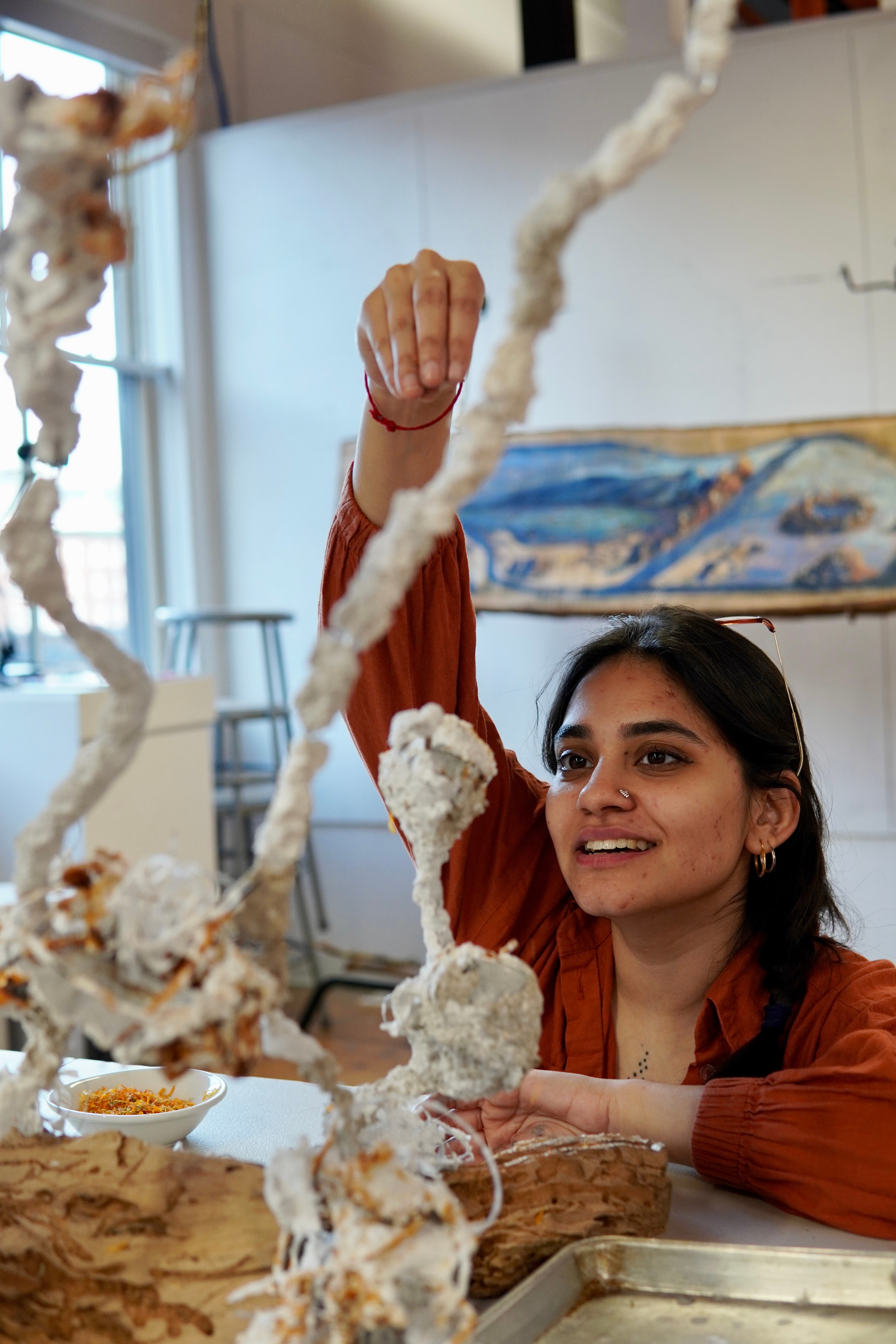 A South Asian woman smiles as she lifts and examines a looping, textured white sculptural form made from paper pulp stuck on a wire armature, rising from a white table surface.