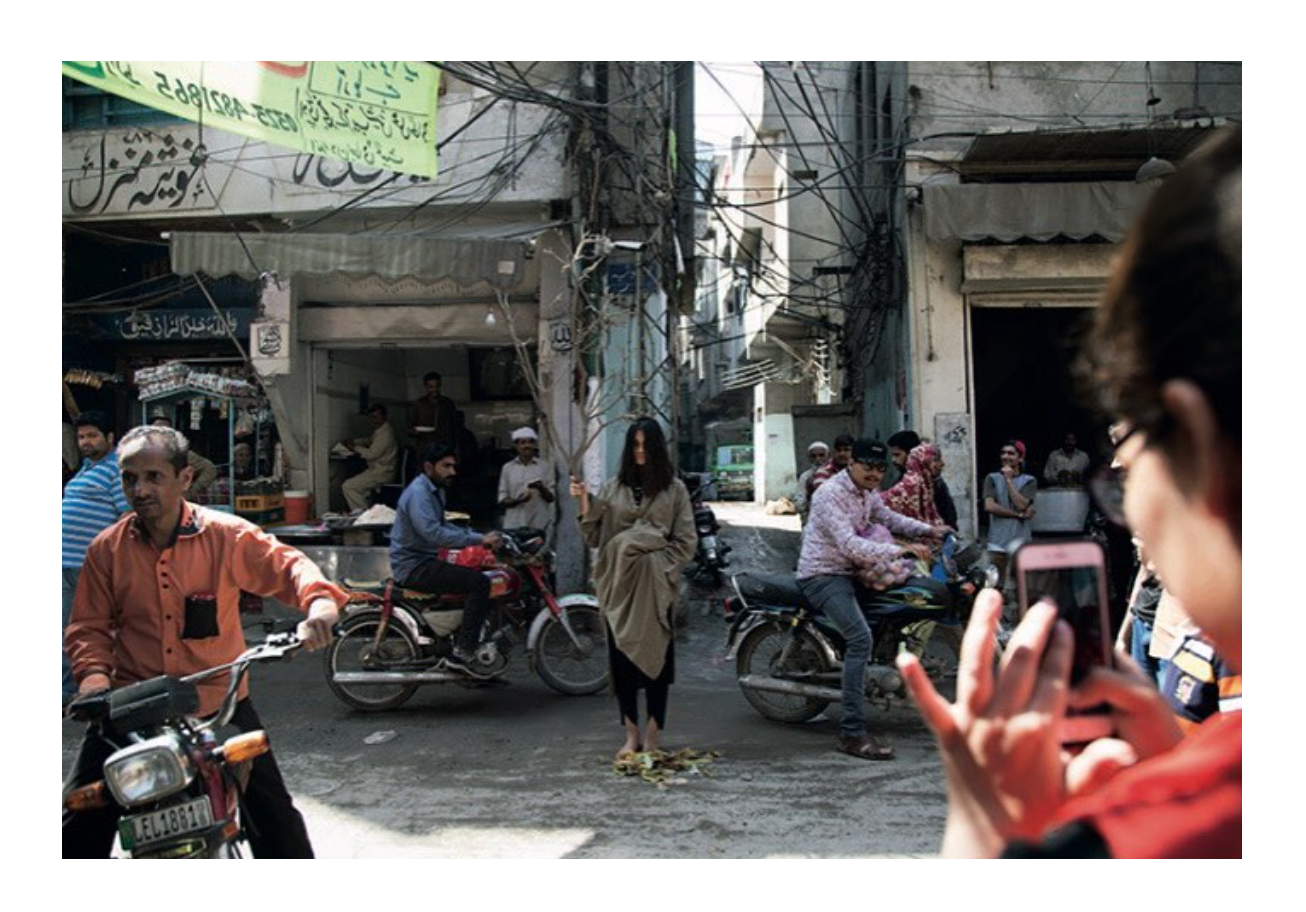A busy urban street in Pakistan, shot from a slight distance. At the centre of the frame, a figure stands still in a khaki draped garment, barefoot, holding a bare tree upright in the middle of traffic. 