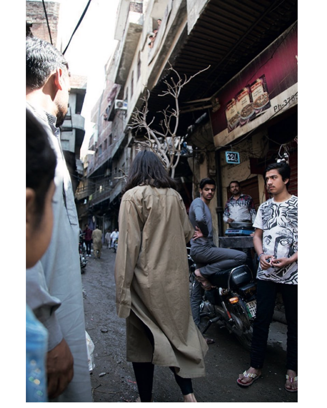 A narrow street in a Pakistani city, photographed from behind. A figure in a long khaki kurta walks barefoot away from the camera into the crowded lane, carrying a bare, branching tree — held upright