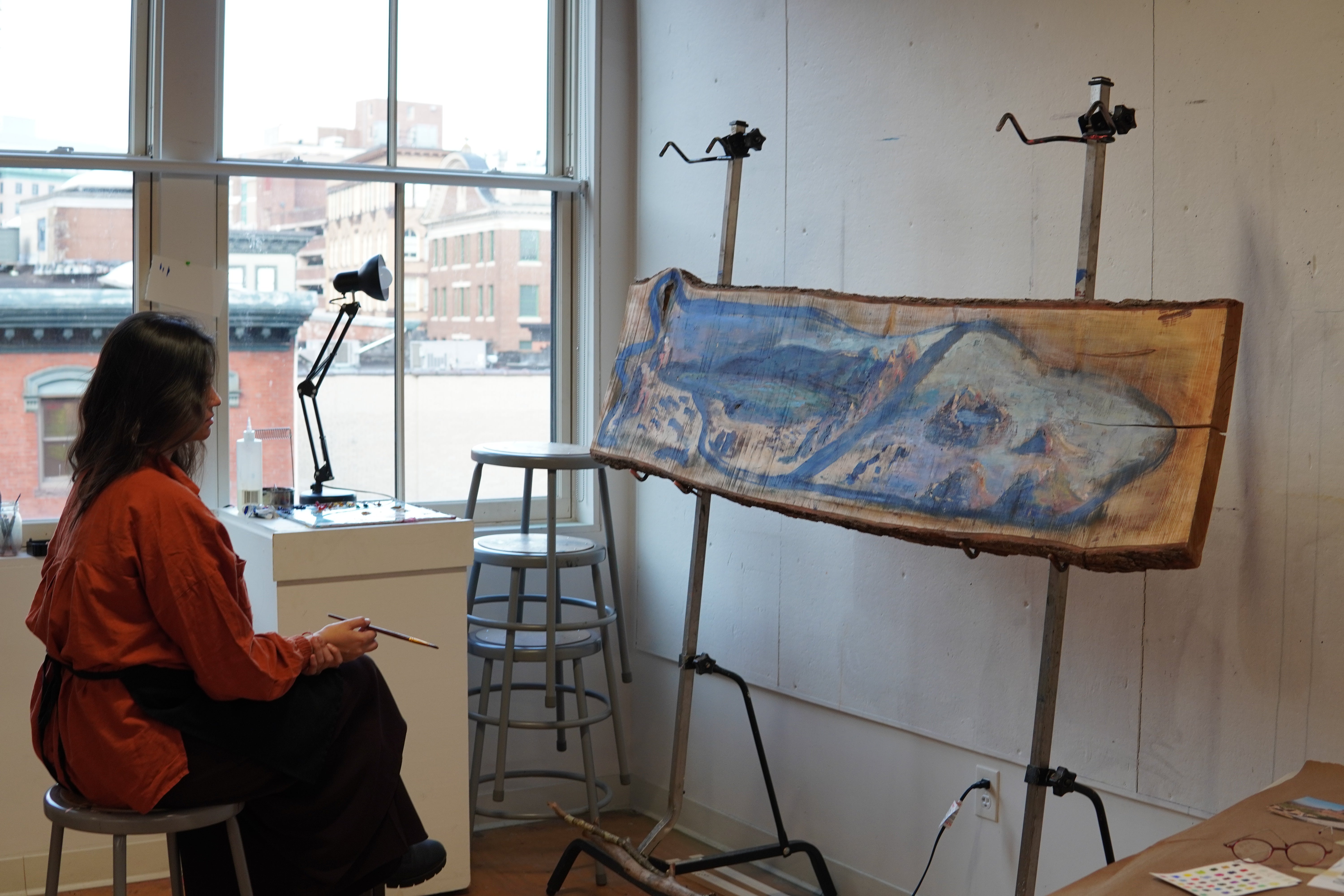 A South Asian woman in a rust-coloured top sits on a metal stool in a high-windowed studio, holding a paintbrush and studying a large live-edge maple panel mounted on a standing easel in front of her.