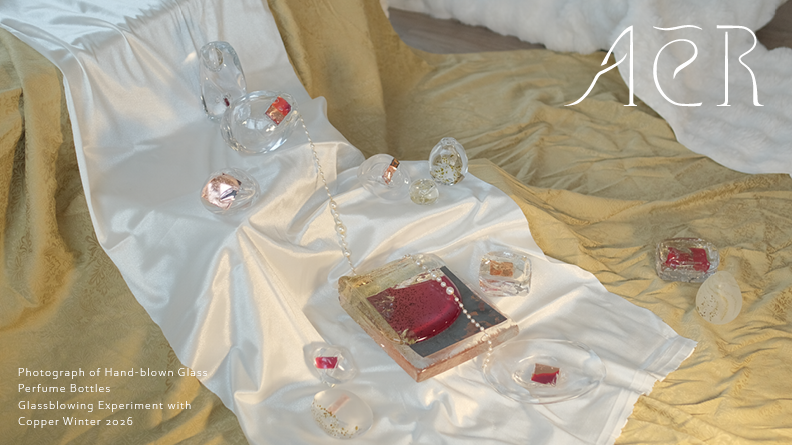 Hand-blown glass perfume bottles arranged on draped white and golden green fabric, each containing small red and copper elements. The composition is softly lit, highlighting transparency, reflections, and material textures.