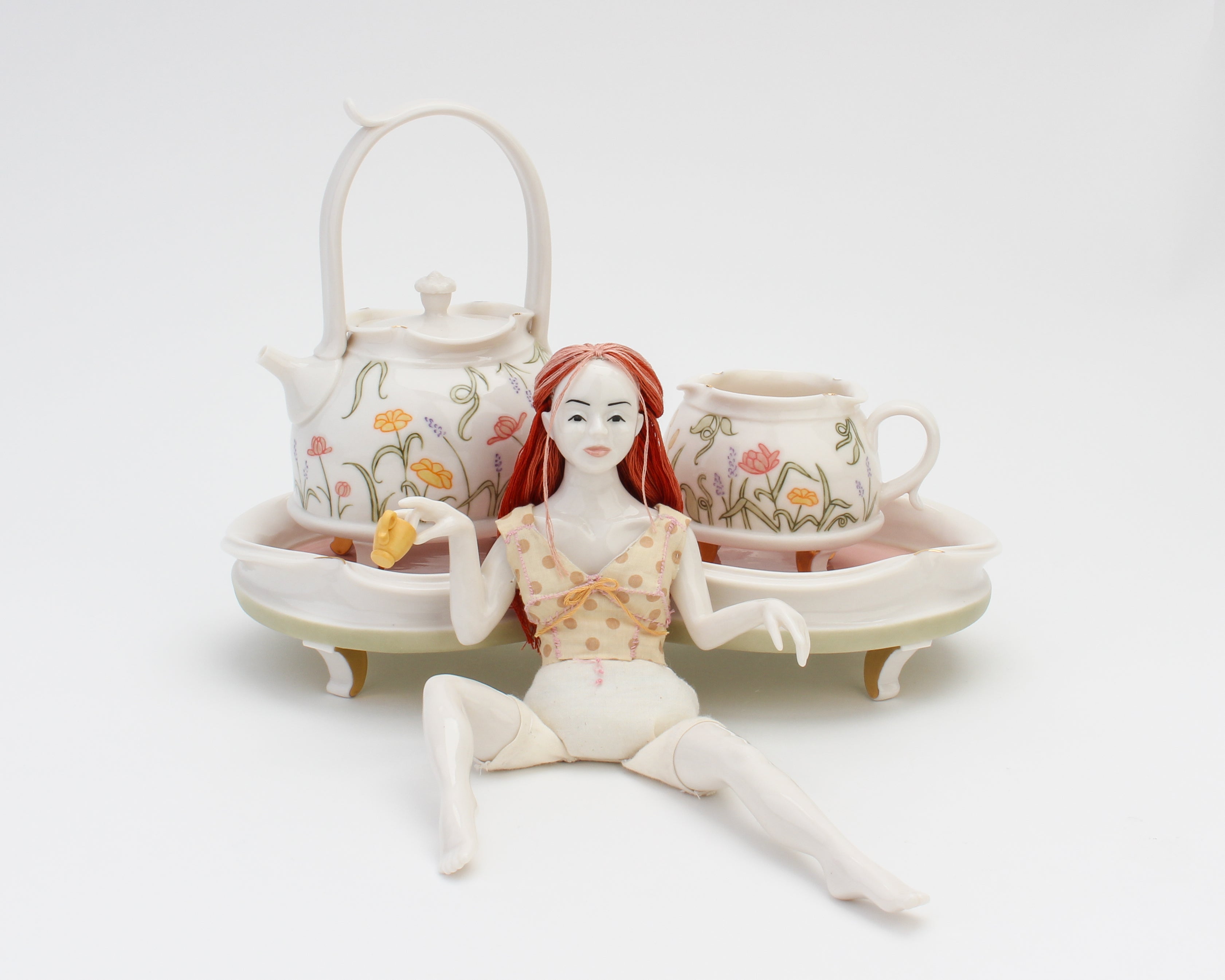 A dainty doll sitting with legs spread apart in front of a human-sized tray, teapot, and teacup decorated with floral designs