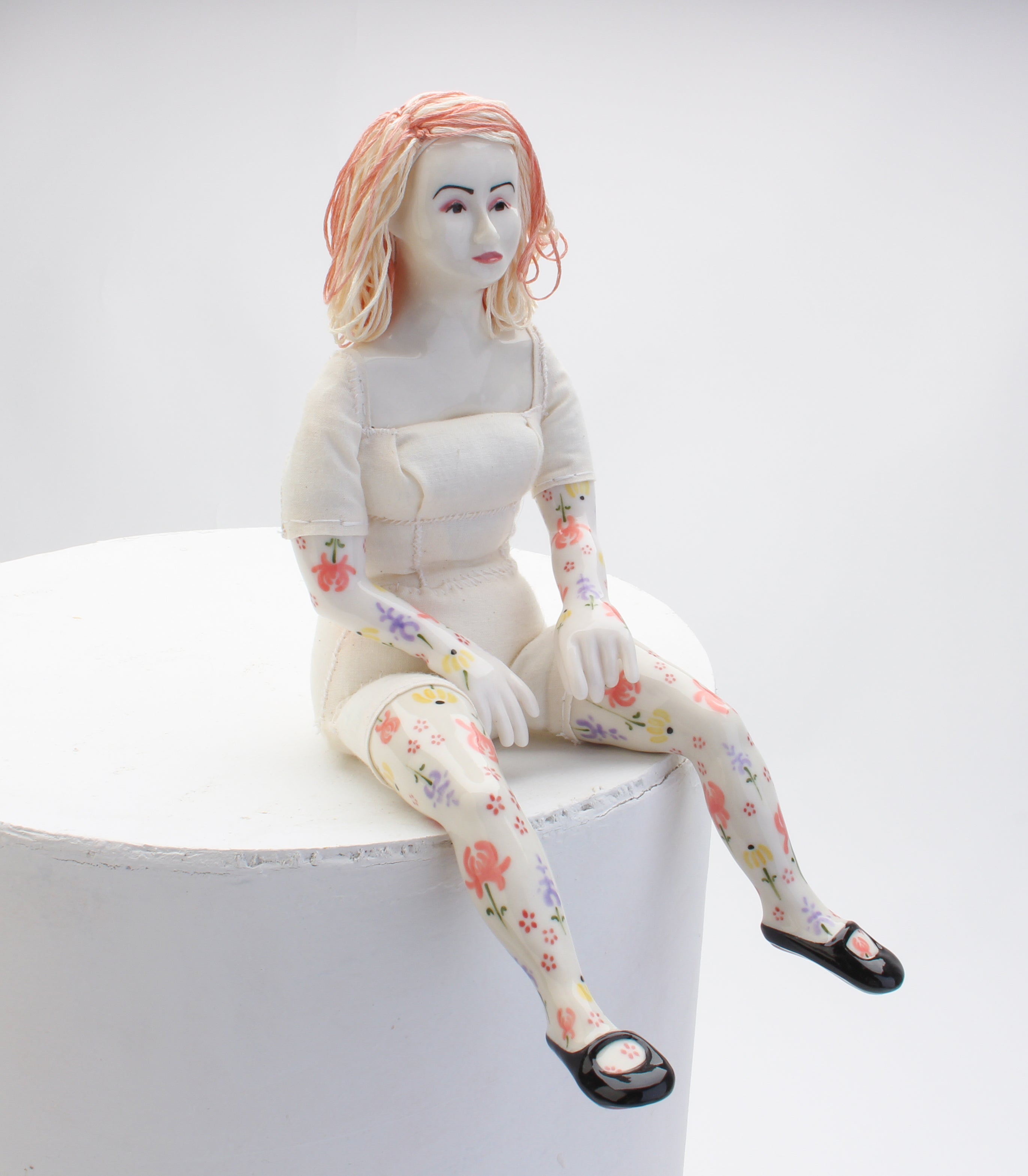 A porcelain doll with white clothes, pink hair, and floral designs on arms and legs sitting on the edge of a pedestal