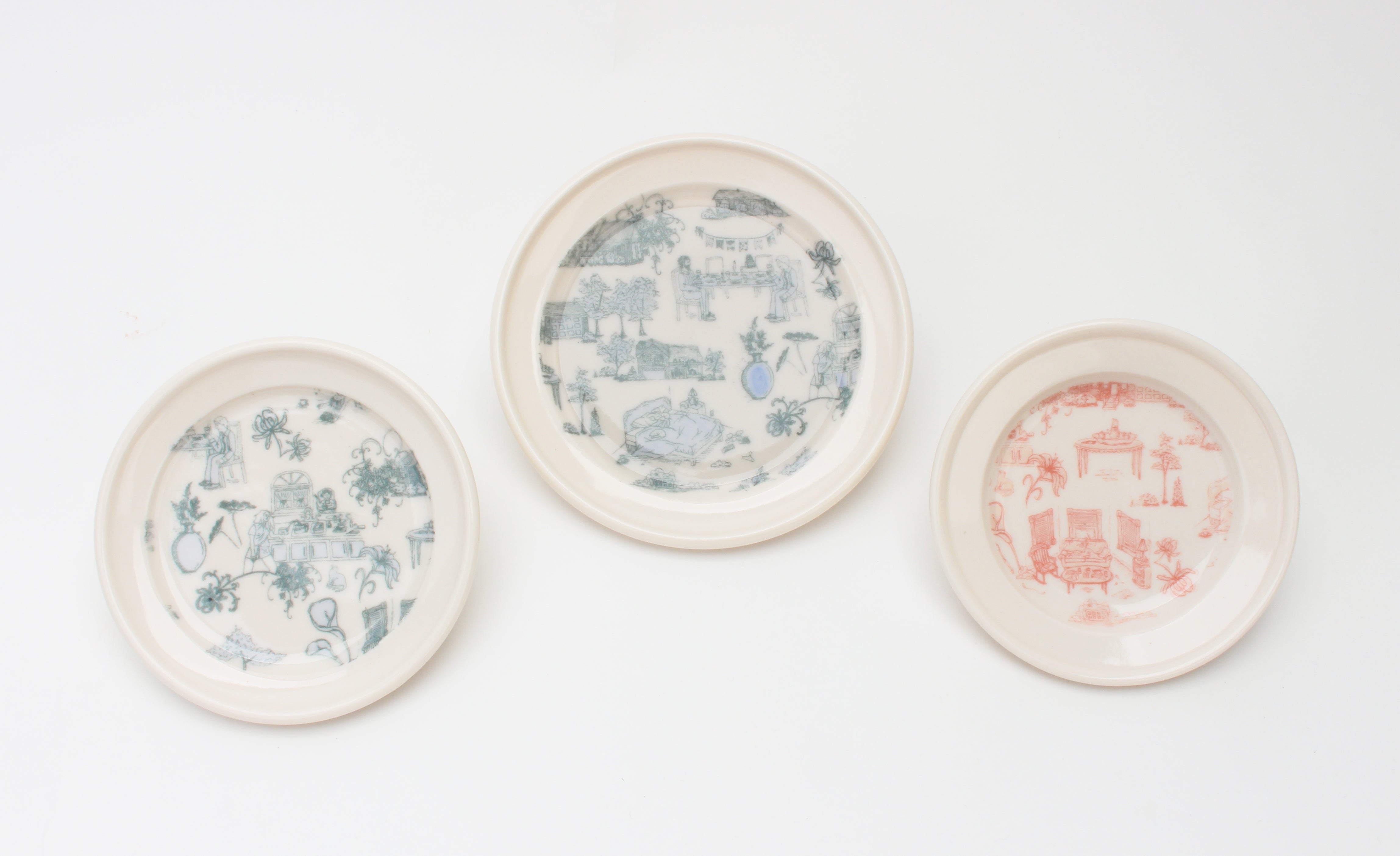 Two white plates with a blue pattern of illustrated cluttered domestic scenes and one white plate with the same pattern in pink