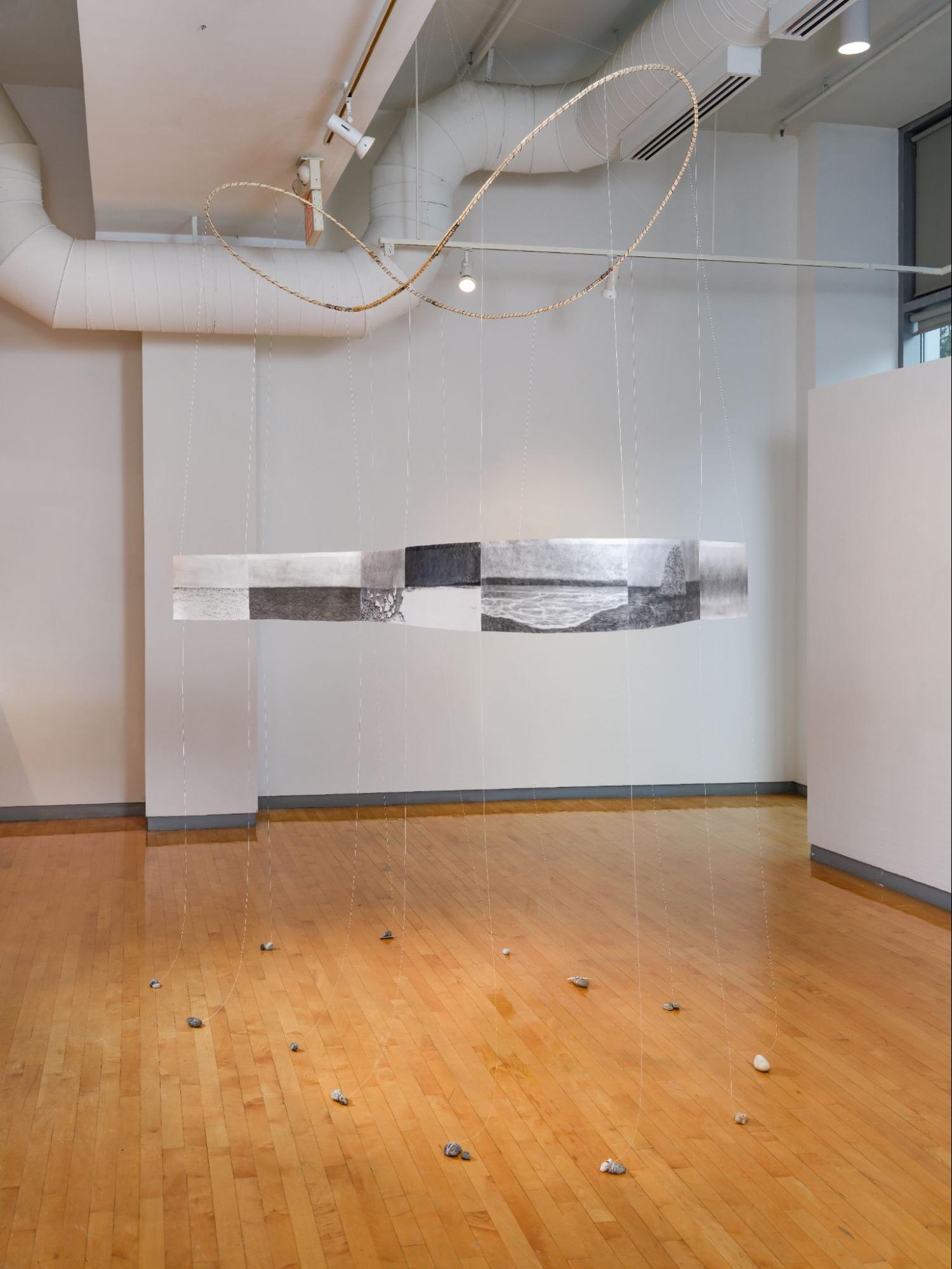 Suspended circular band of drawings hung by thin strings, with stones on the floor beneath in a gallery space