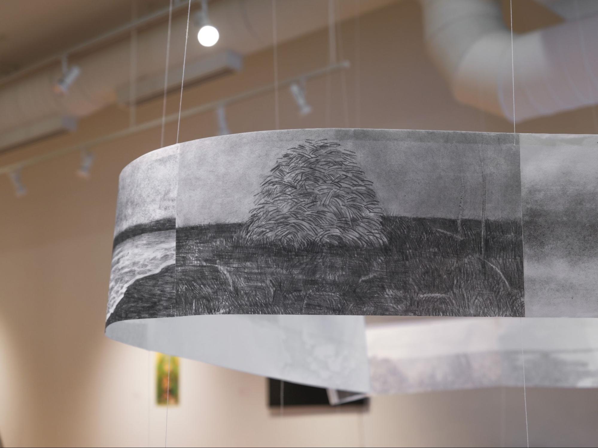 Close-up of a curved paper strip with charcoal landscape drawing, suspended by thin vertical strings