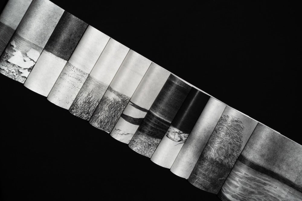 Accordion-folded paper strip with charcoal landscape drawings forming a continuous horizon against a black background