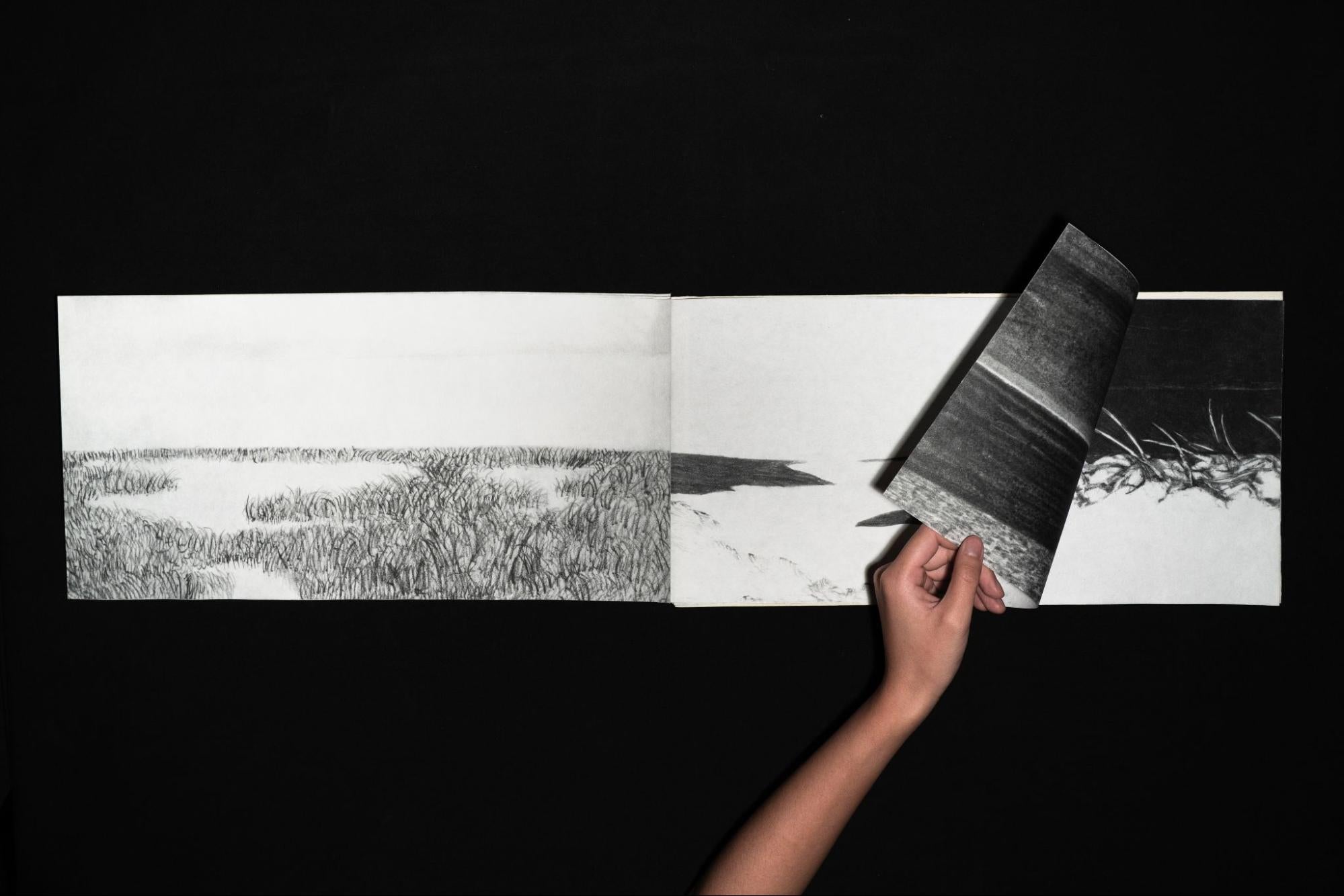 Hand lifting a page of an accordion book revealing a continuous charcoal landscape drawing across connected panels