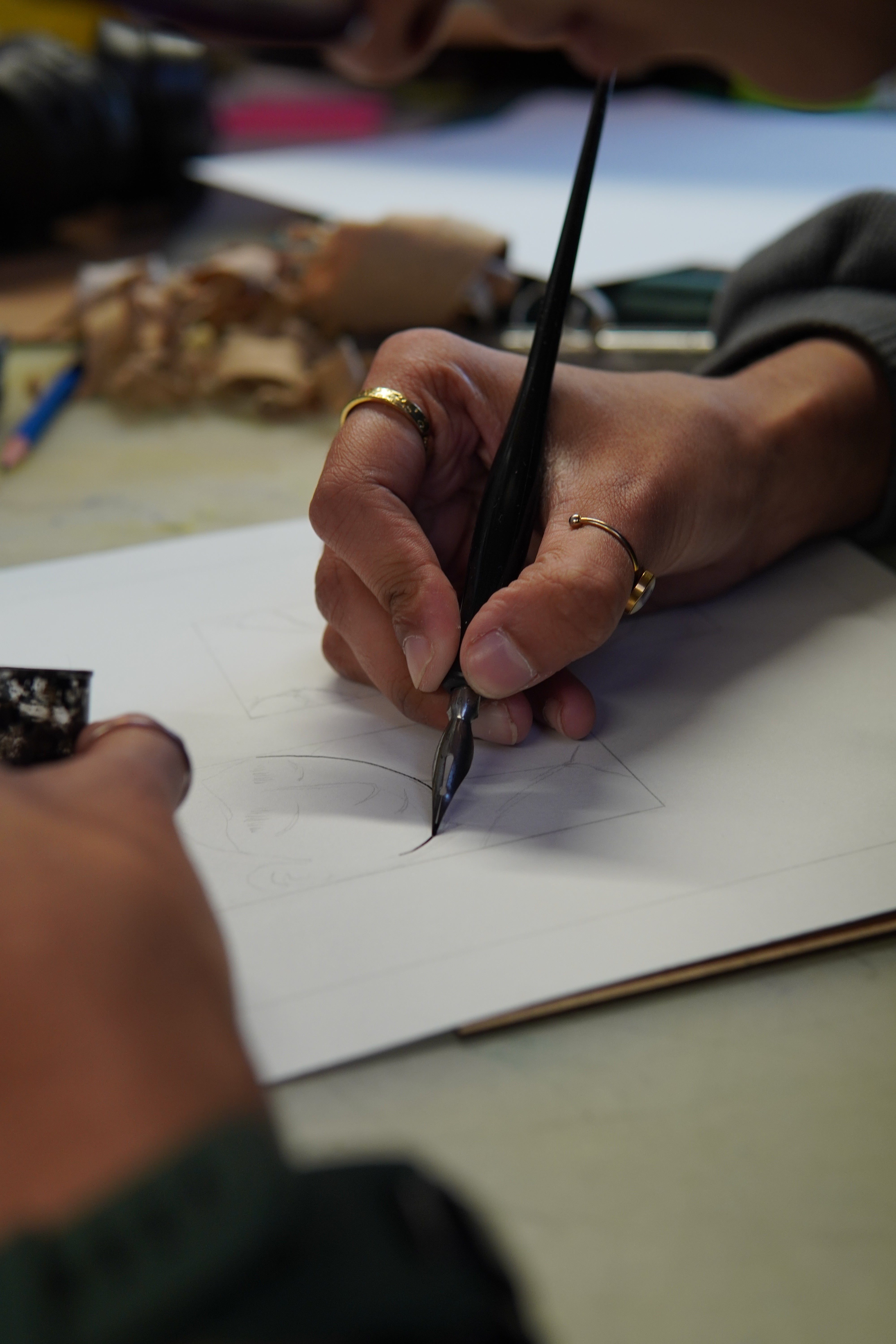 Image shows artist inking a hand-drawn comic page on paper, applying ink over pencil lines with a dip pen as part of the final linework stage of comic production.