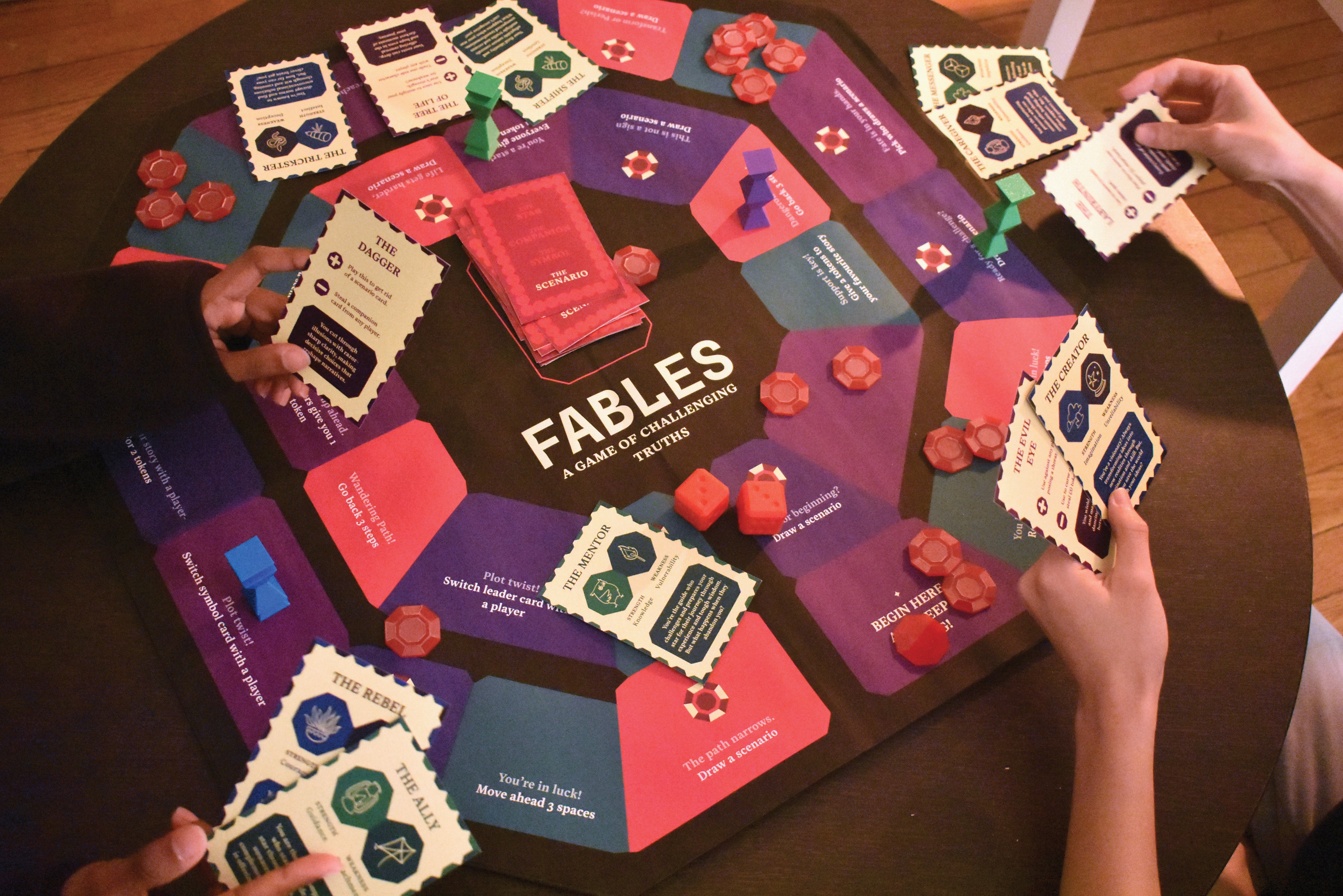 A colorful board game layout displays "Fables: A Game of Challenging Truths" with character archetype cards, scenario cards in red and purple, coral-colored tokens, and red dice, arranged on a wooden table with players' hands holding cards.