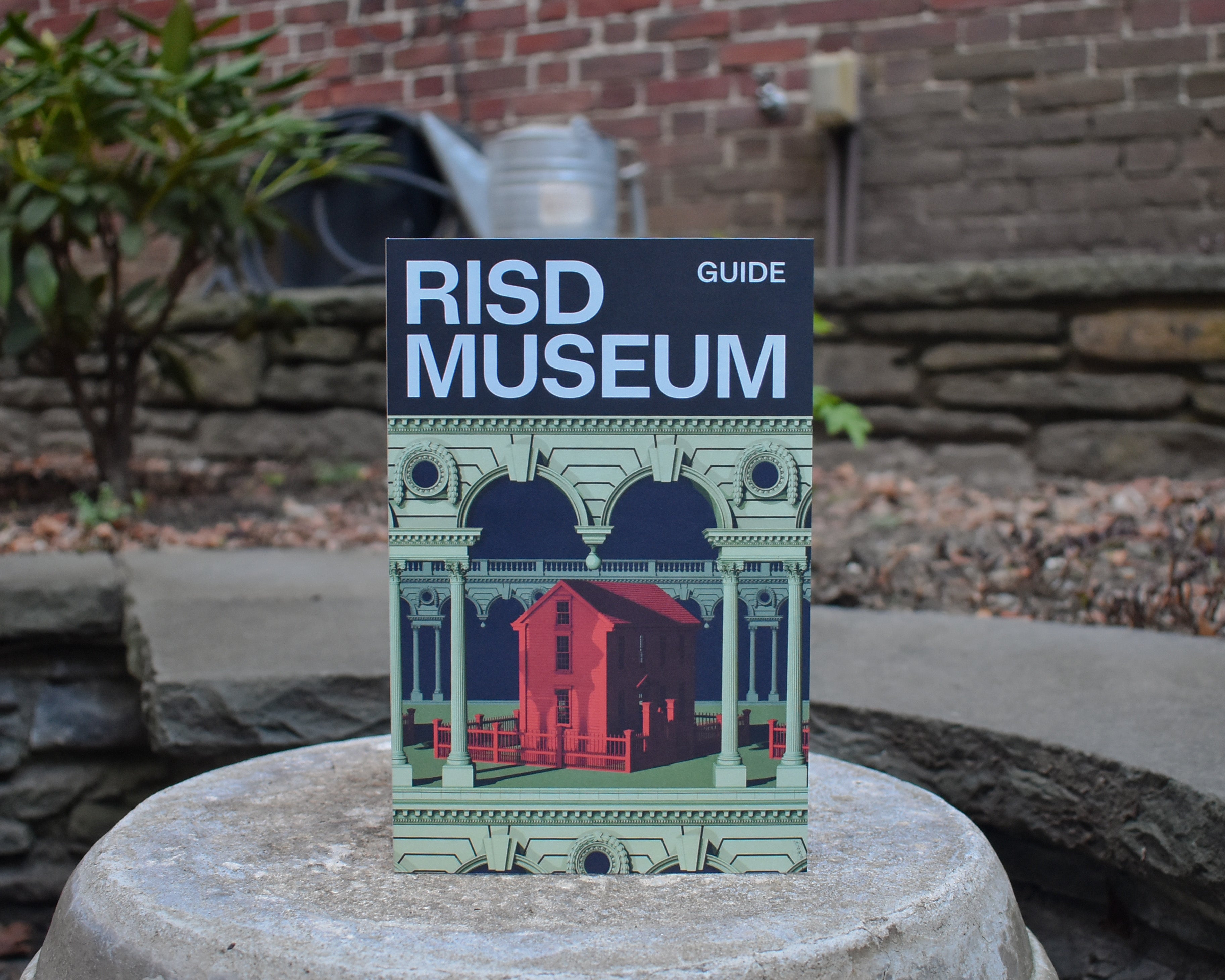 A RISD Museum Guide cover stands upright on a stone surface, featuring a navy blue header with light blue typography and an illustration of the museum's red brick architecture framed by ornate green classical detailing.