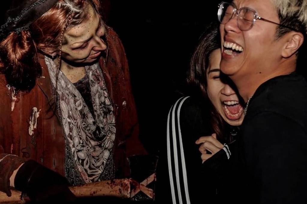 Three people are visible in the dark at a haunted attraction park. One is a scare actor in a scary costume. The other two, visitors to the attraction, huddle together in response to the scare.
