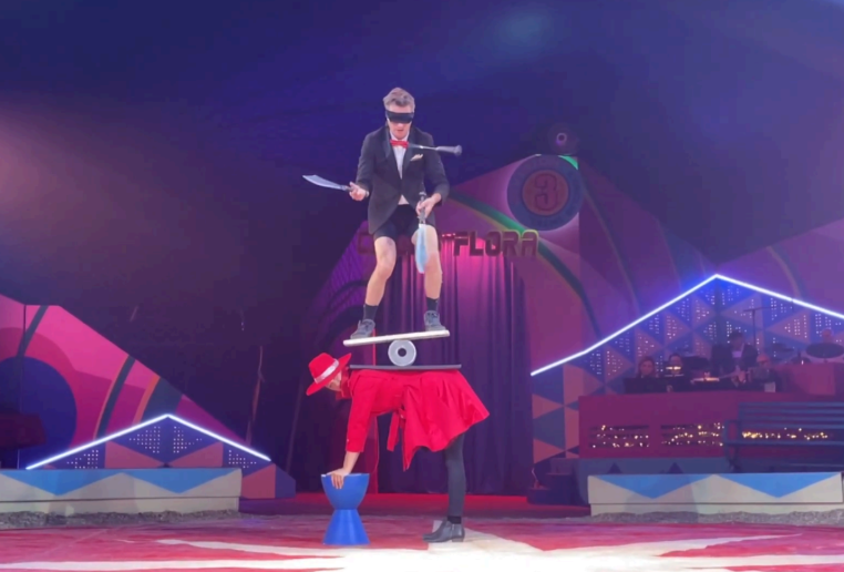 A scene from a circus performance. A woman is bent over to support the juggling act happening on her back. Balanced on her back, a blindfolded man stands on a moving apparatus.