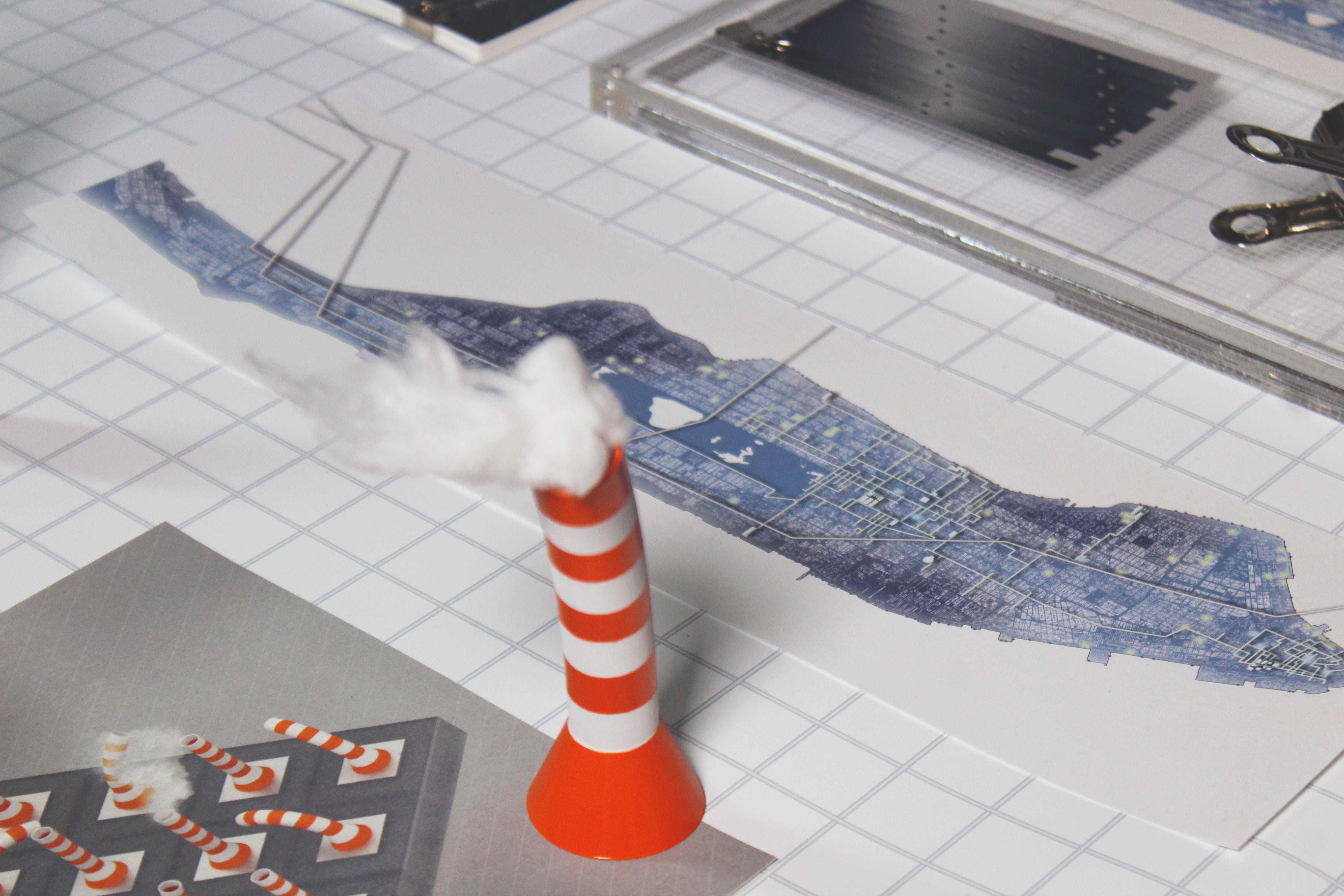 Photograph of an orange-and-white steam stack in the foreground, and a map of Manhattan in the background. The map portrays water systems within the island, identifying supply, points of control, and escape.  