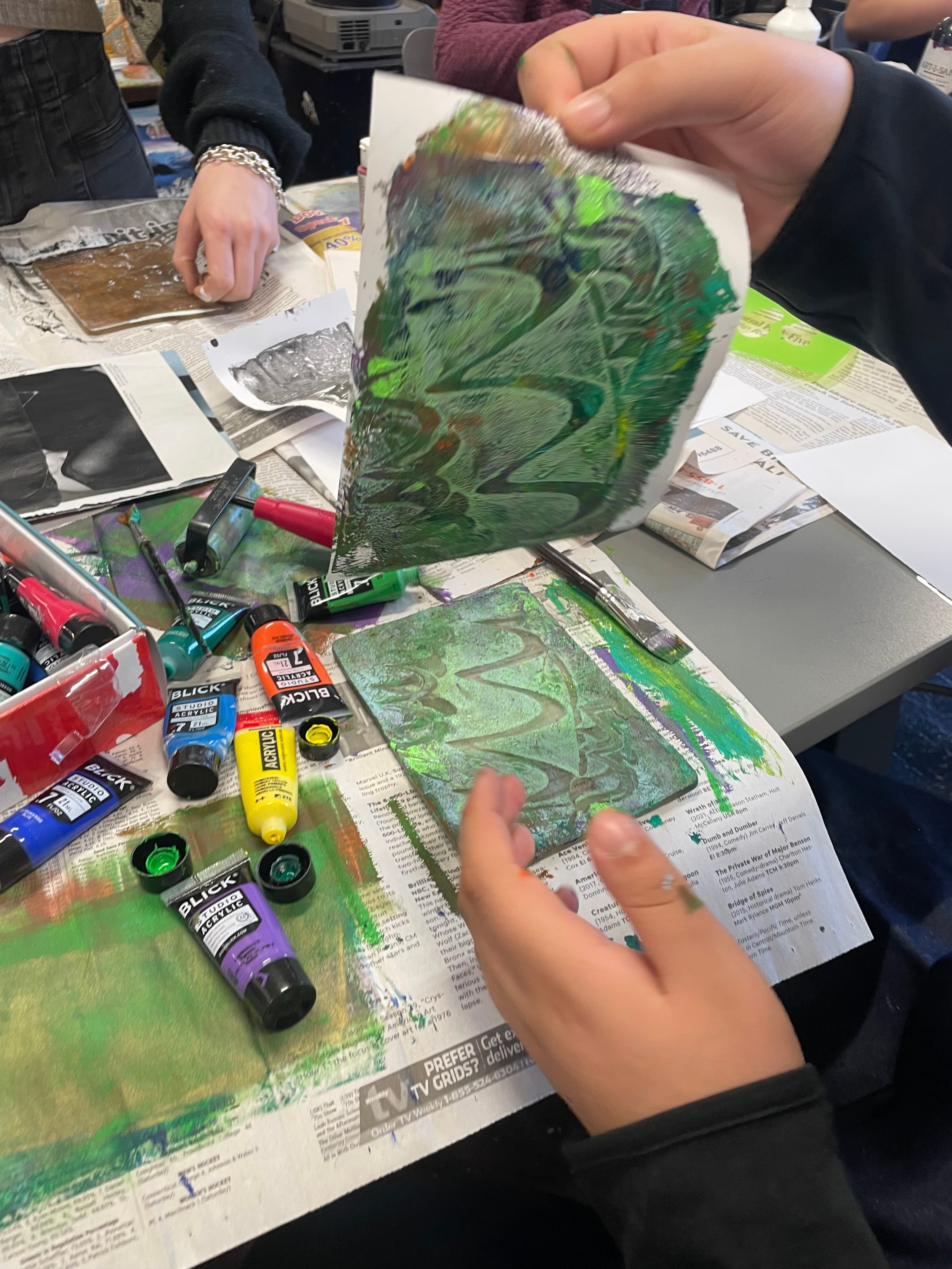 student lifting green abstract image on paper from a geli plate on top of a table covered in paint tubes and newspaper.