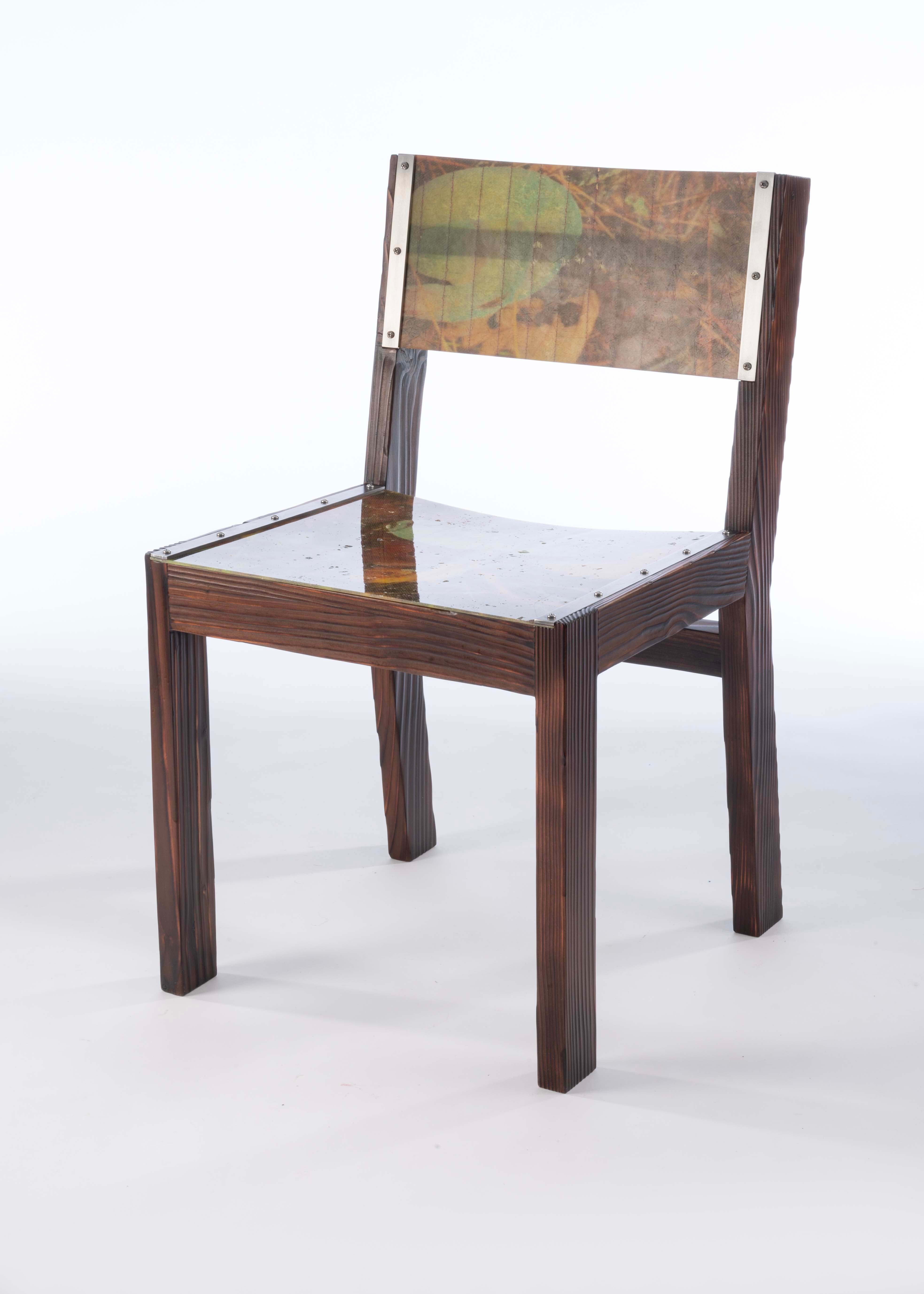 A chair with a blocky frame made of darkened burnt wood with a seat and back panel made from fiberglass that has images of the forest floor laminated inside.