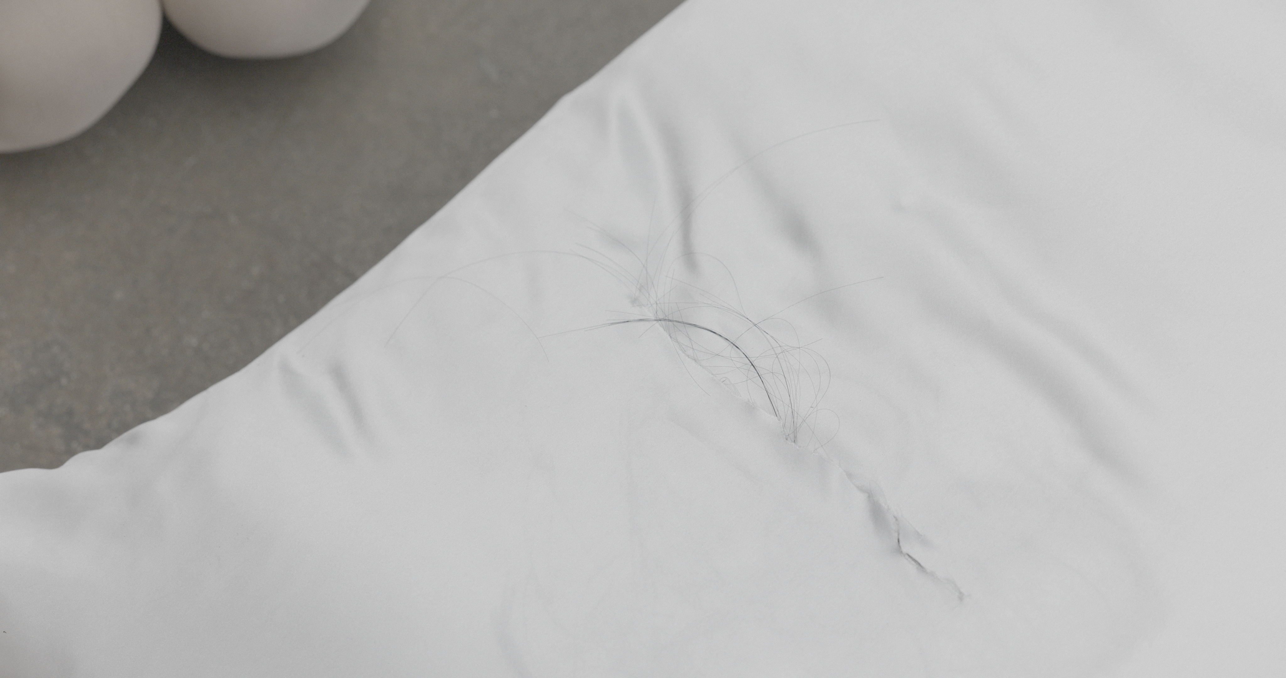 Close view of a white pillow on concrete floor with a small tear revealing strands of dark hair emerging from inside; smooth fabric contrasts with rough ground.