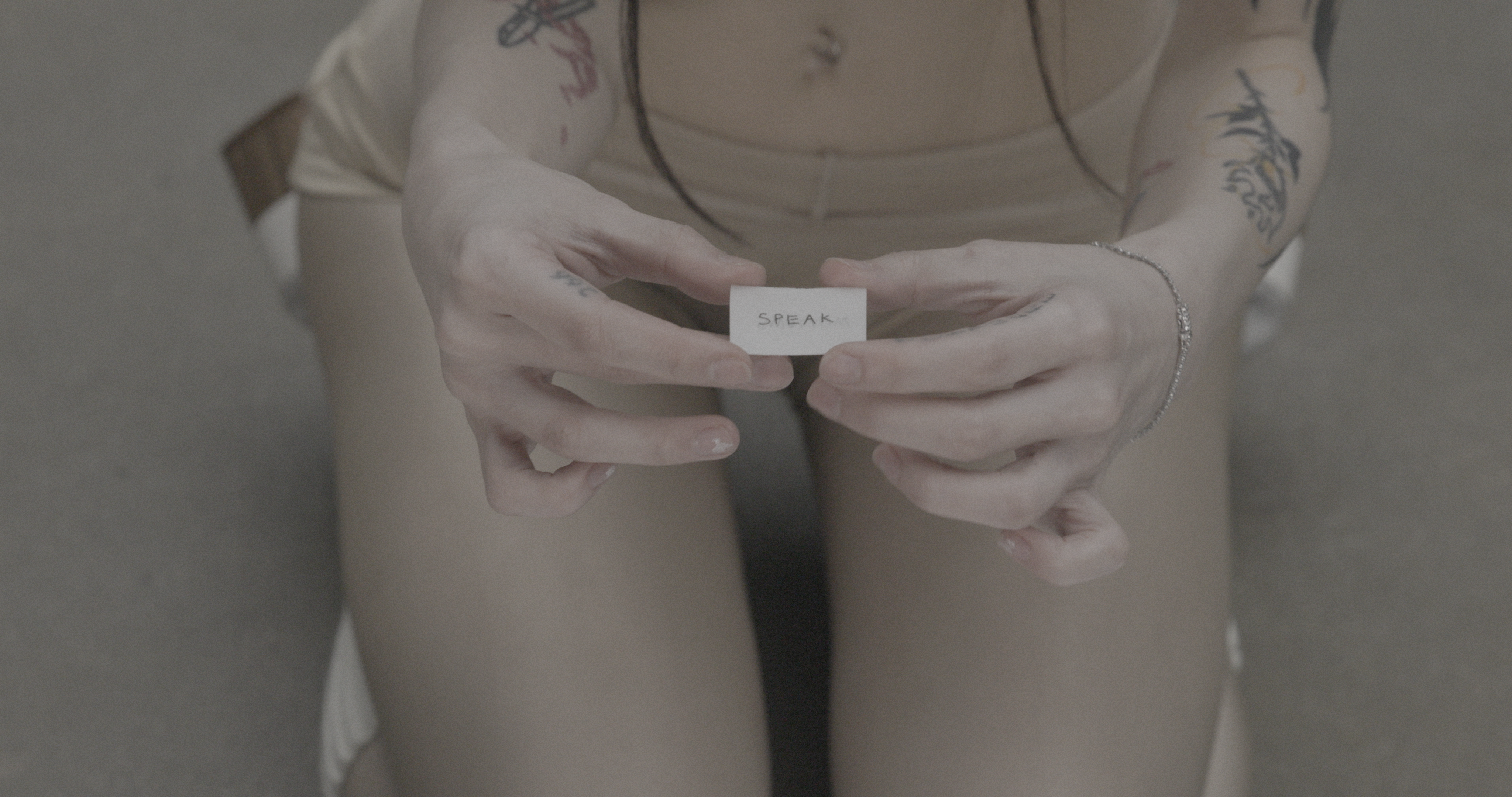 Hands hold a small white paper labeled “SPEAK” between fingers over nude-clad thighs; seated body visible, soft lighting, muted tones, tattoos faintly visible on arms.