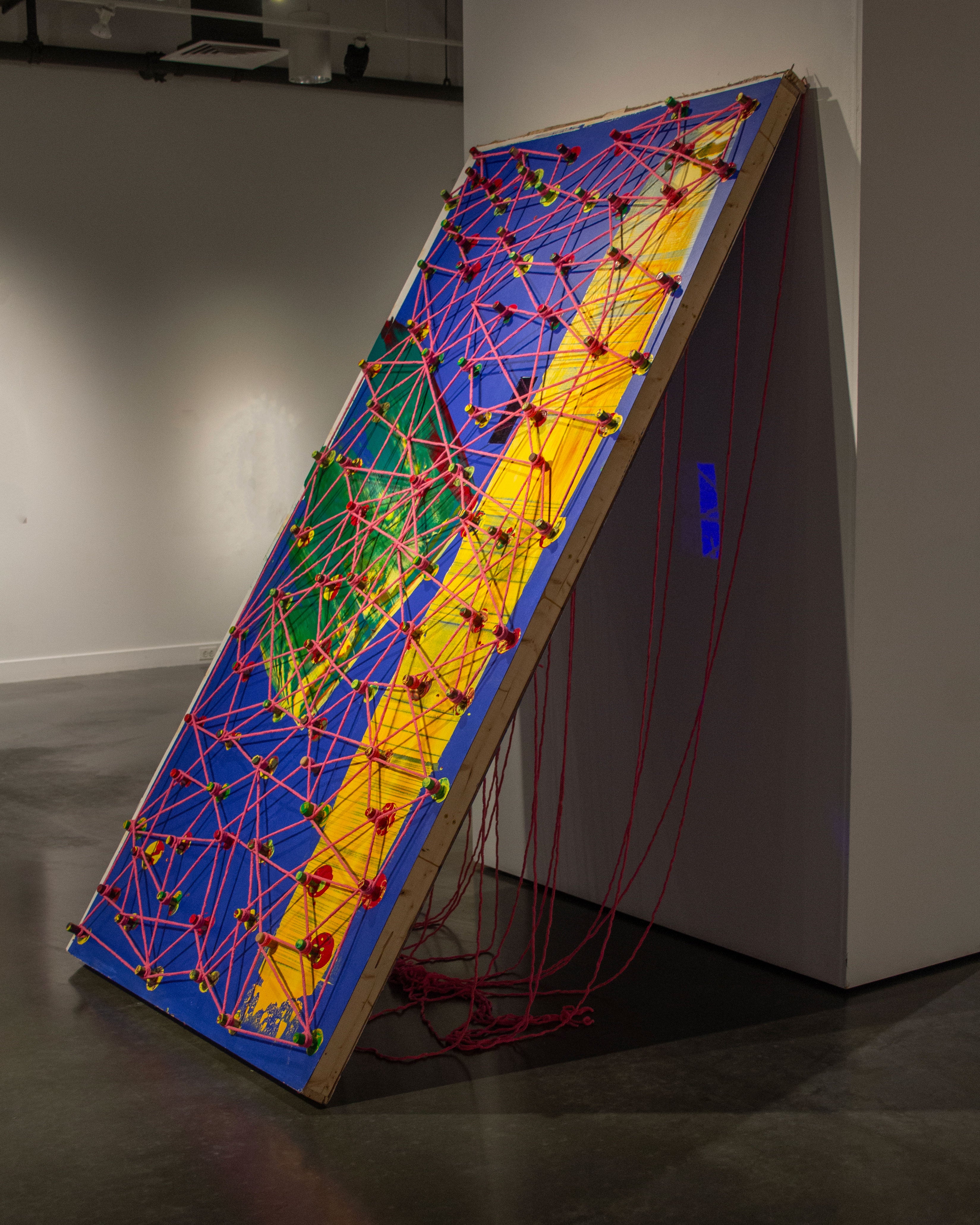 This represents a big blue wooden panel, stand diagonally against the wall. It has long yellow streak, with wine many corks. all the wine corks are connected through a pink yarn, represents an abstracted language.