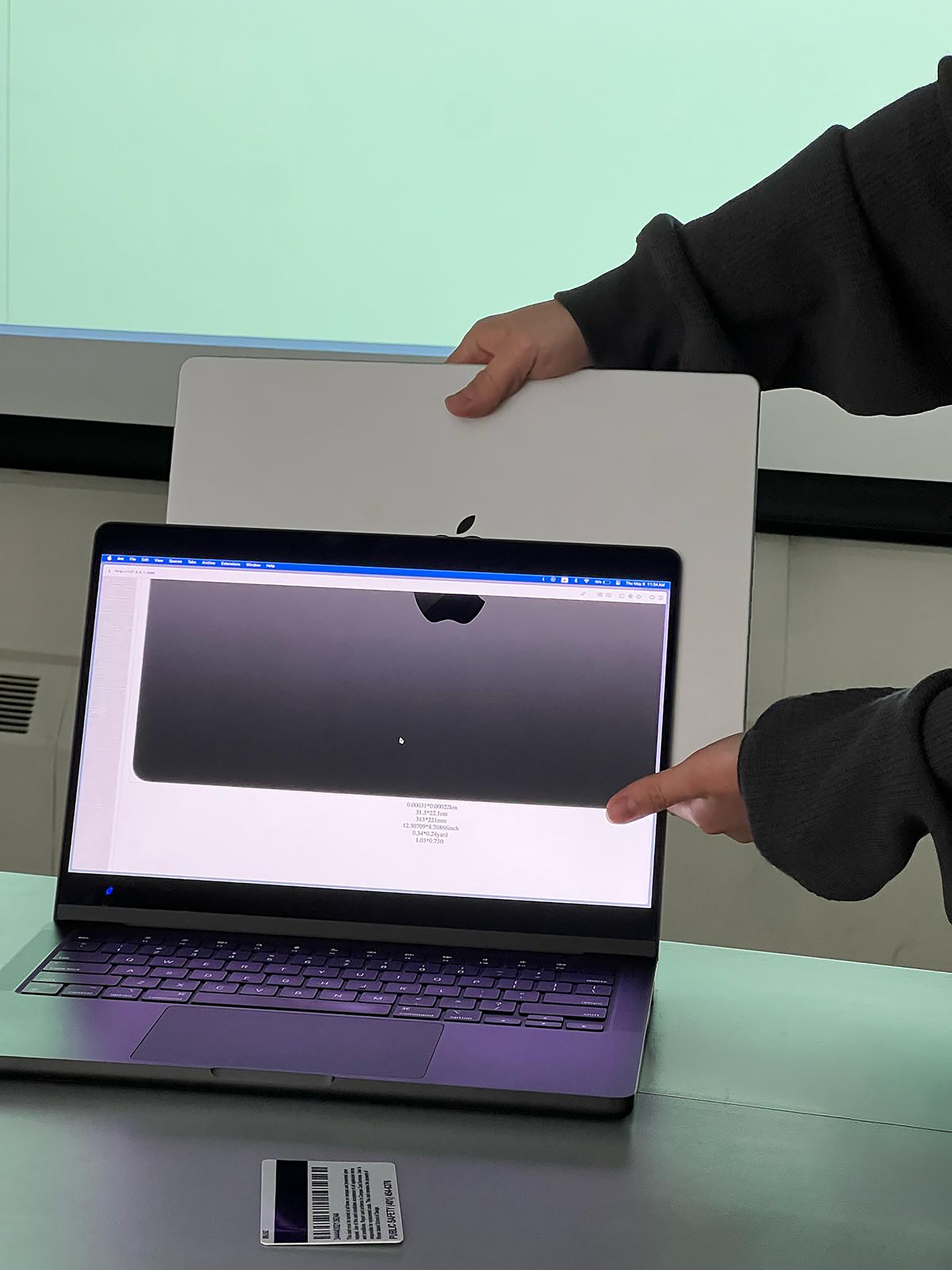 Two hands hold a closed silver laptop behind an open laptop displaying a grayscale scan of its back. A barcode card rests on the table in front of the screens.