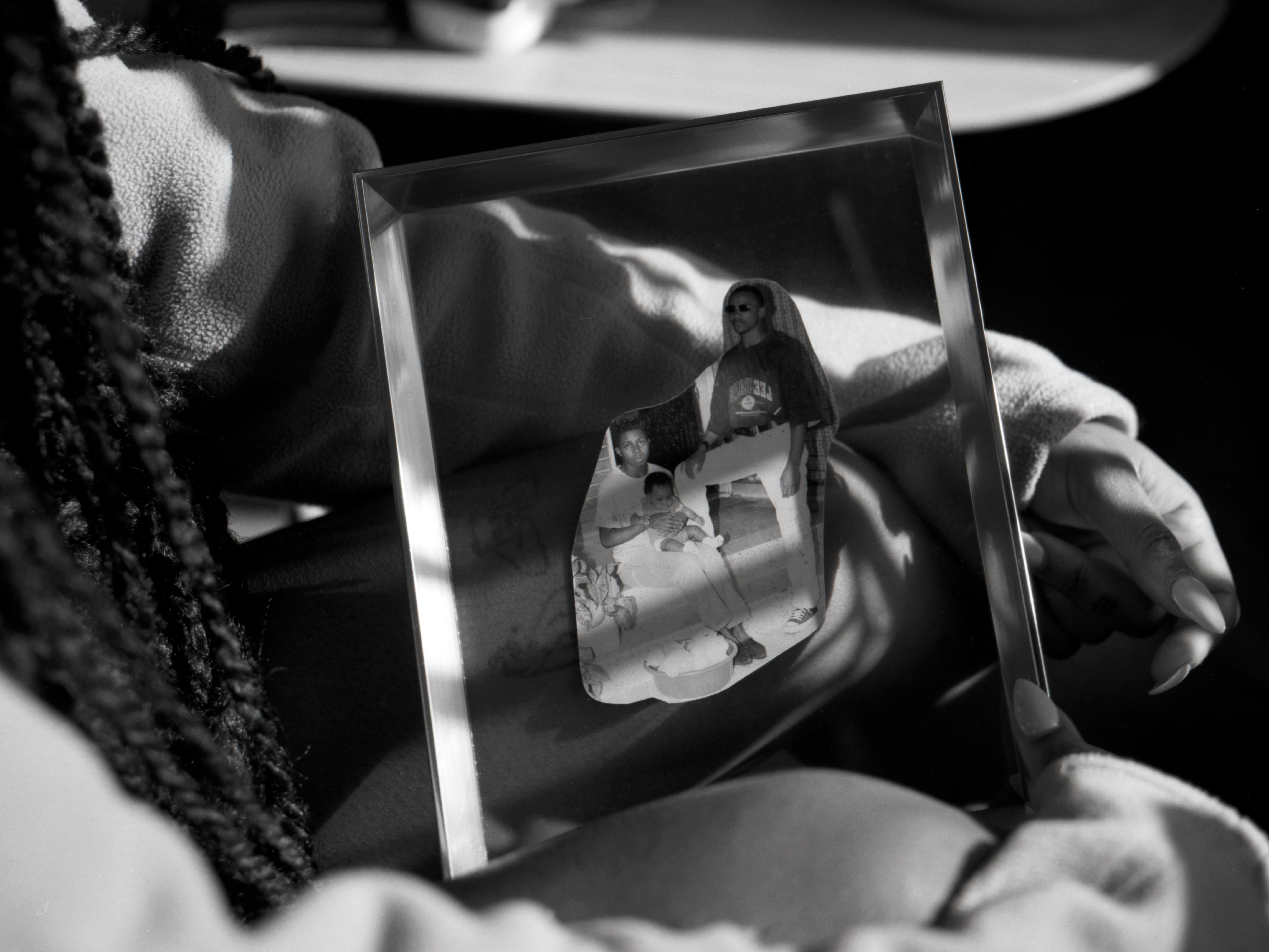 Black and white photograph of a person holding a framed photograph with a torn collage of people in casual attire. There are three figures. Sunlight casts shadows across frame.