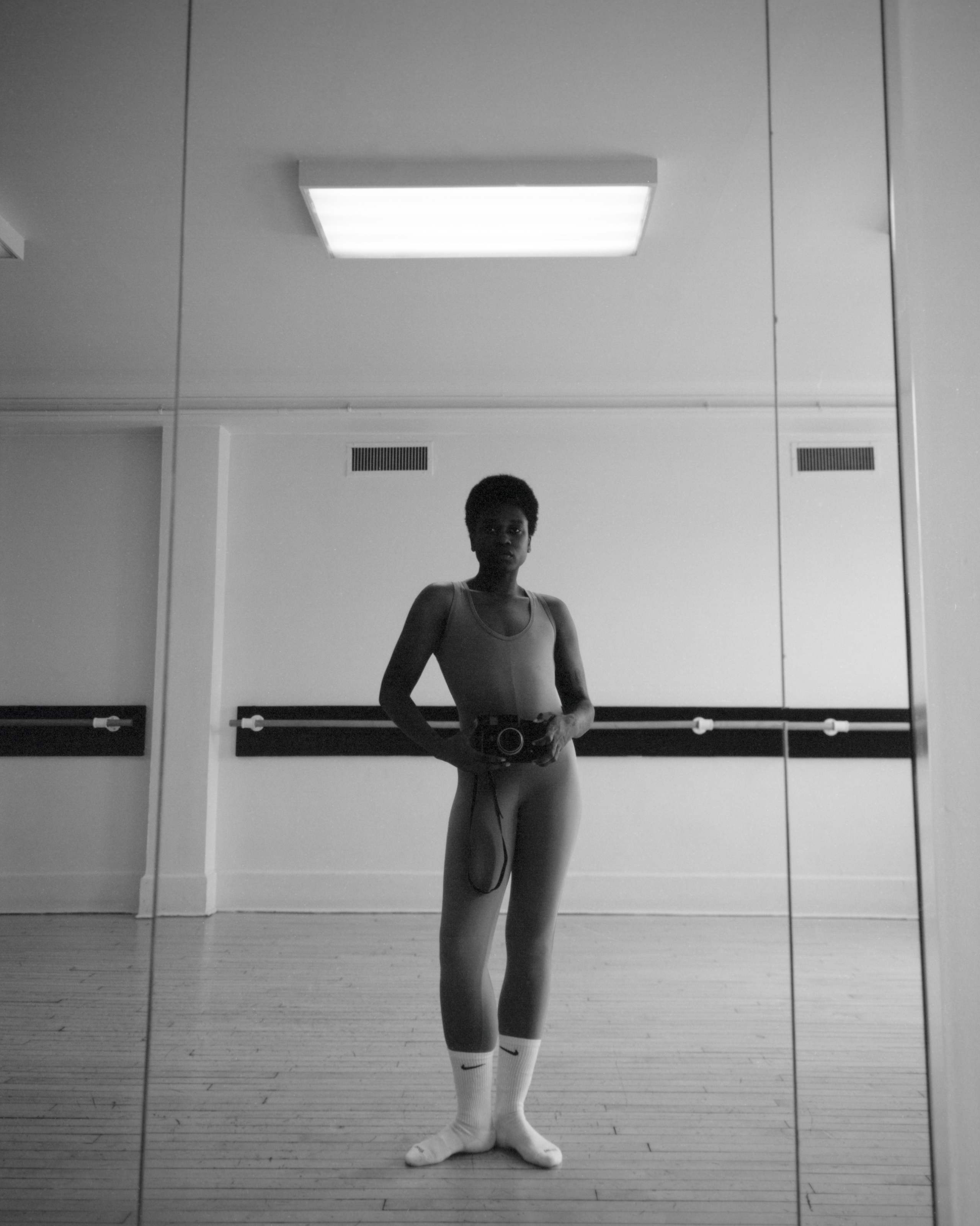 Black and white photograph of a person in a leotard and socks, holding a camera in a mirrored dance studio. The space is minimal and reflective.