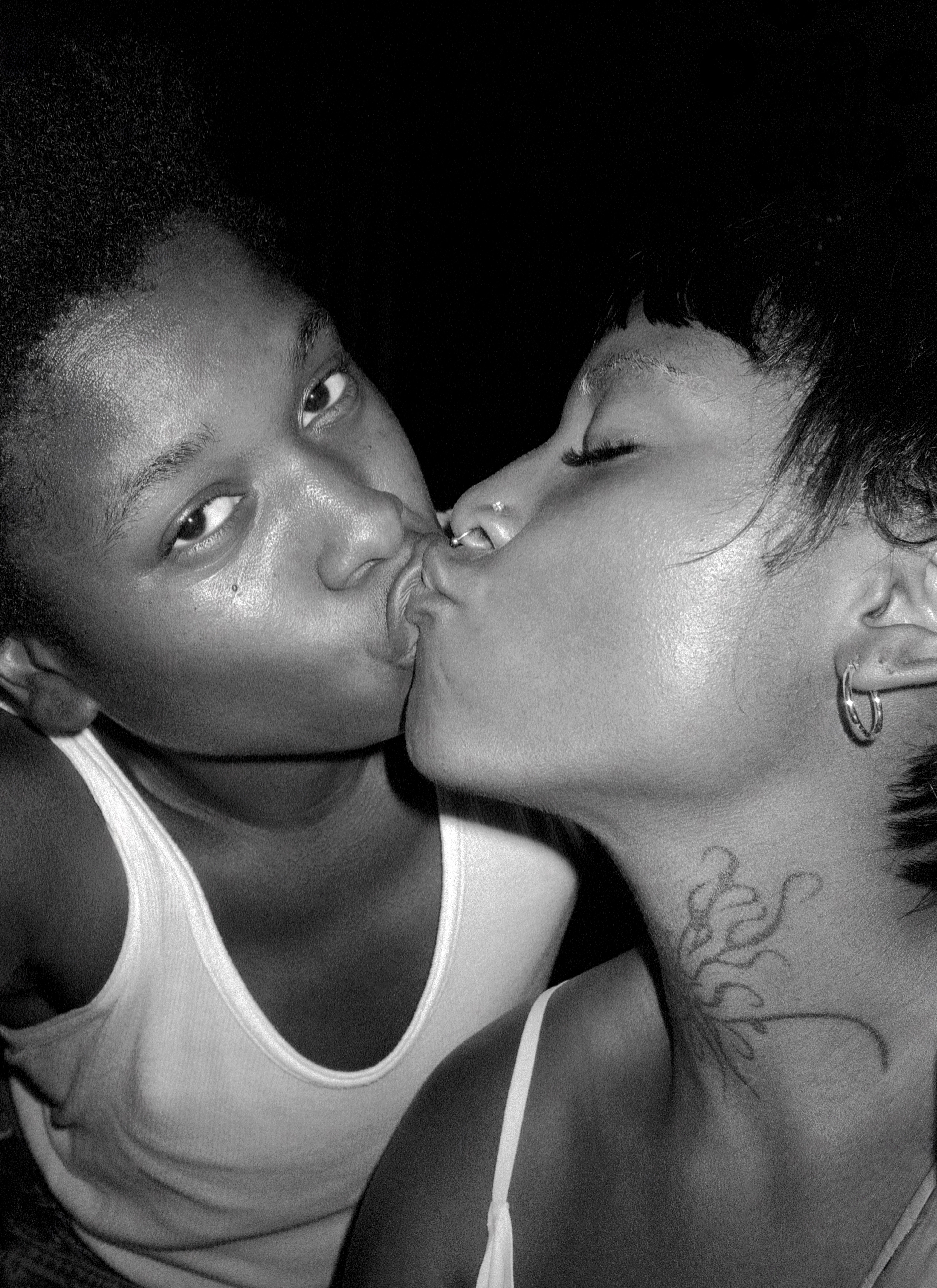 Black and white photograph of two people kiss intimately, their faces close. Person on the left is looking directly into the camera and person on the right has a tattoo on their neck, wearing hoop earrings.