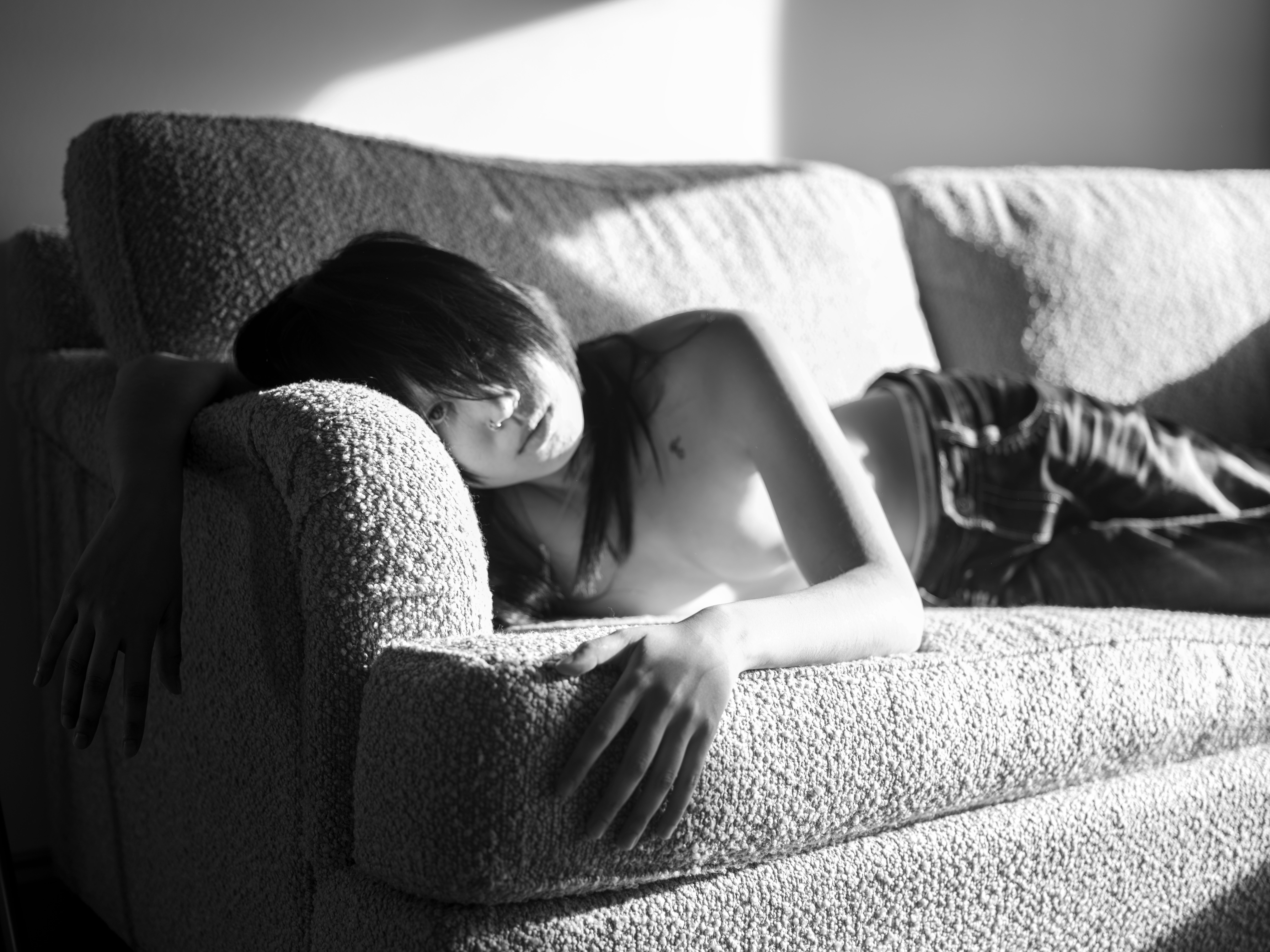 Black and white photograph of a person with long hair lounging on a textured sofa in soft sunlight. Their eyes are almost fully covered from their bangs.