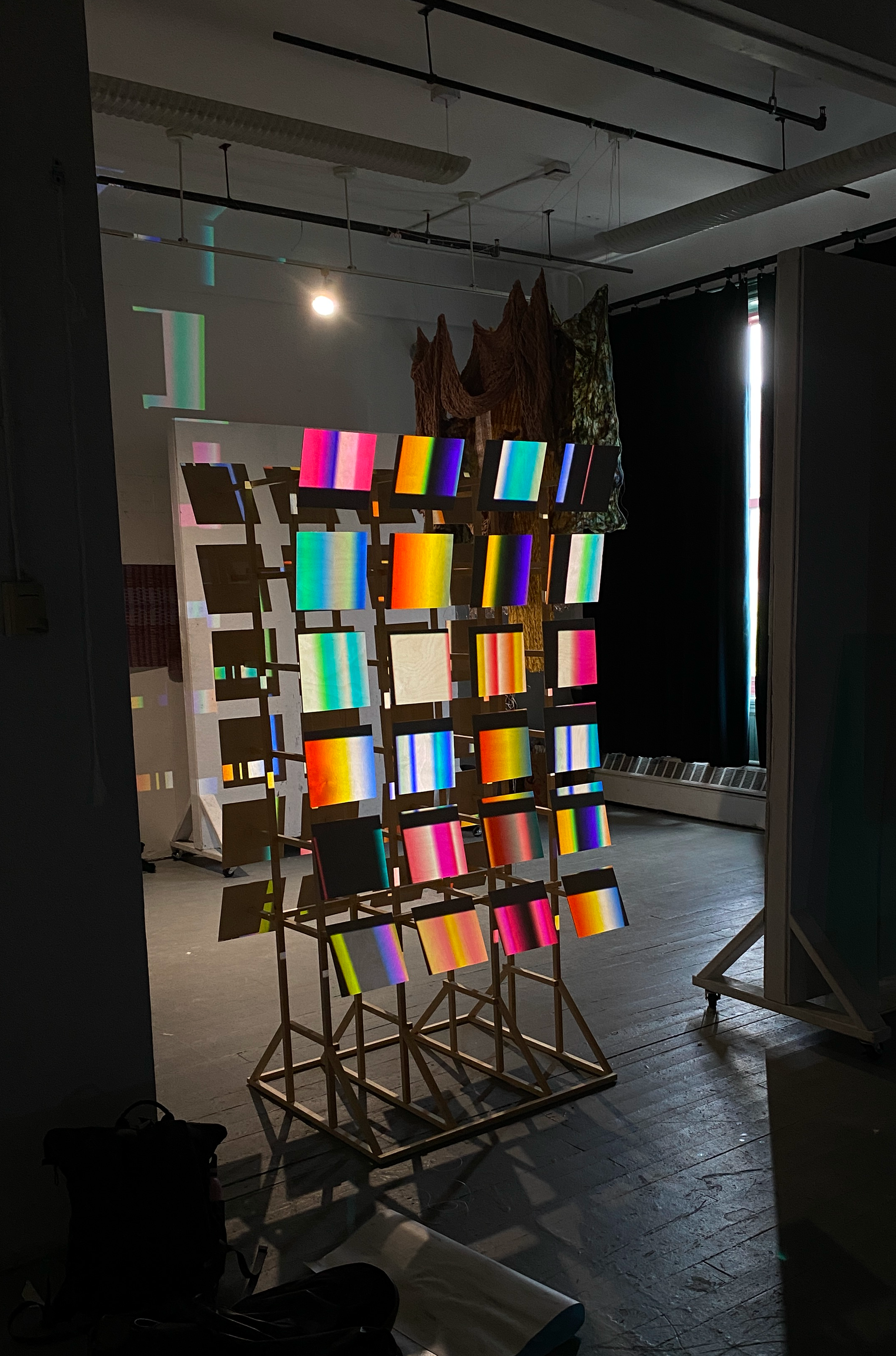 Dim installation view of a wooden structure holding many glowing gradient panels, with projected color bands and shadows filling a studio space.