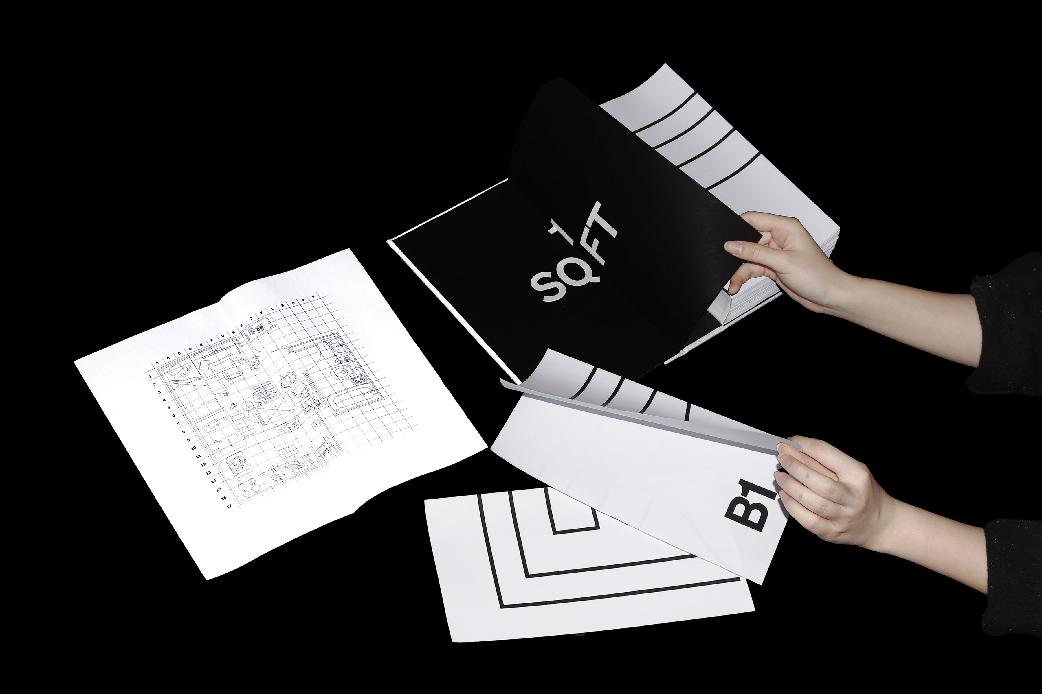 Hands unfold a large black-and-white book-object, revealing spreads with floor-plan grids, thick contour lines, and large room labels against a black studio background.