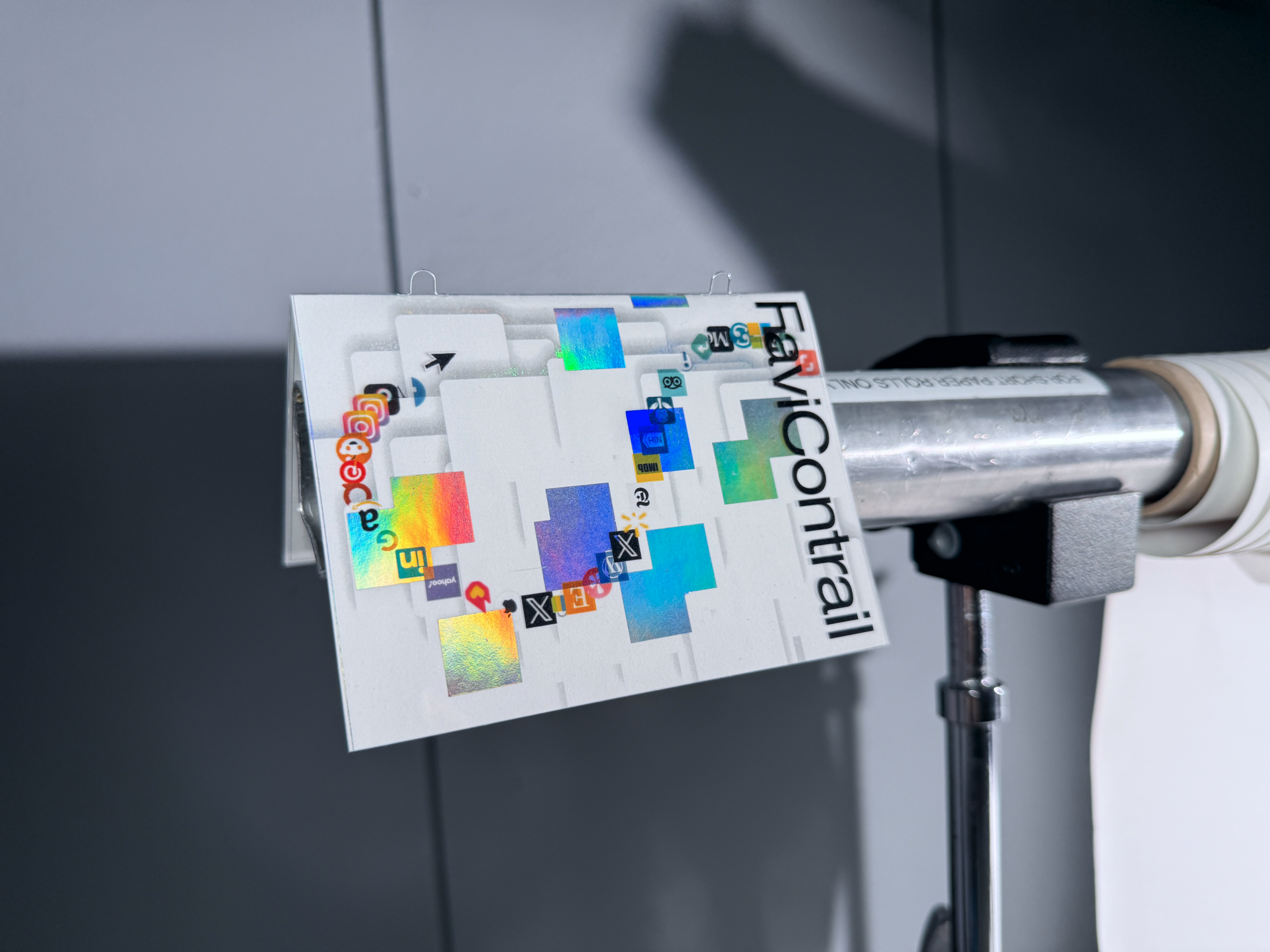 Close-up of the FaviContrail brochure print clipped to a metal pole, showing browser favicons trailing across layered white shapes and holographic color patches.