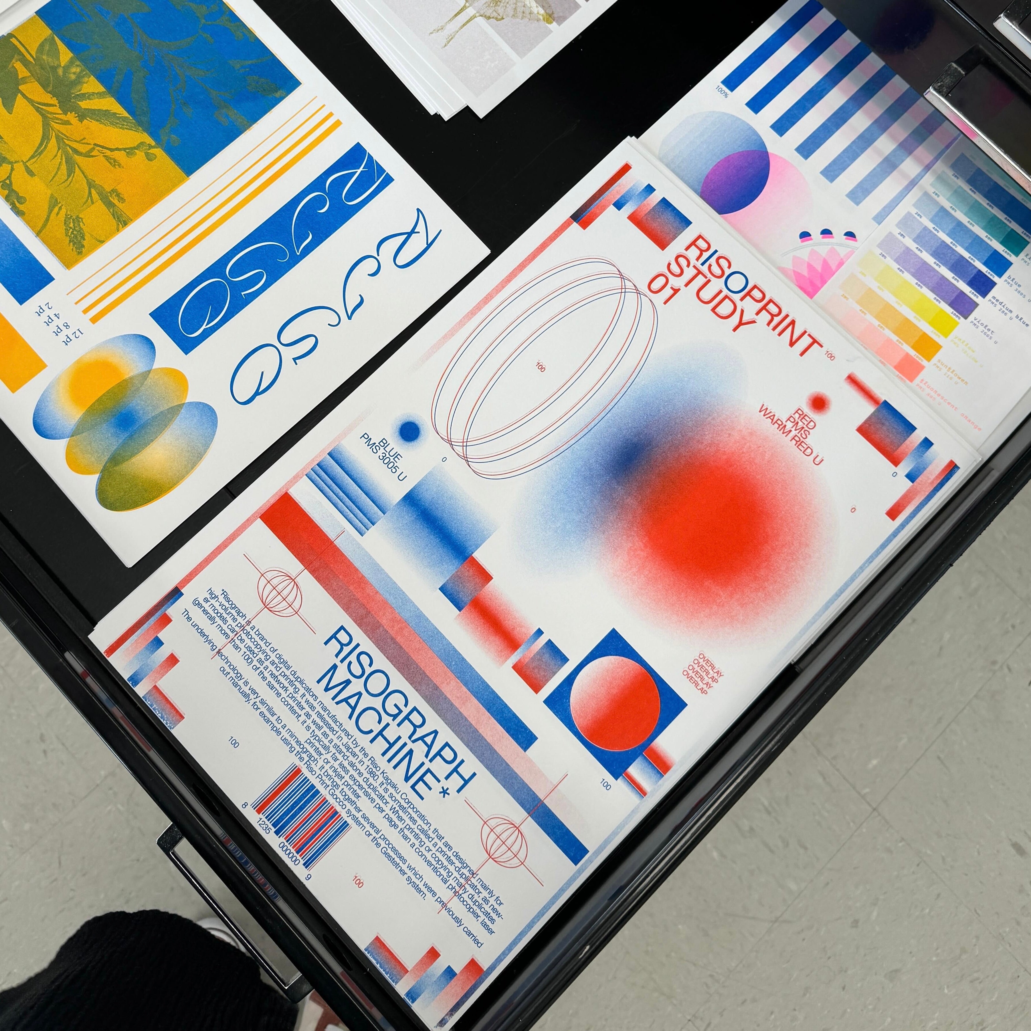 Overhead view of riso-printed poster titled "Risograph Machine" on a table, showing layered blue, red, yellow, and pink gradients, type experiments, color swatches, and geometric forms.
