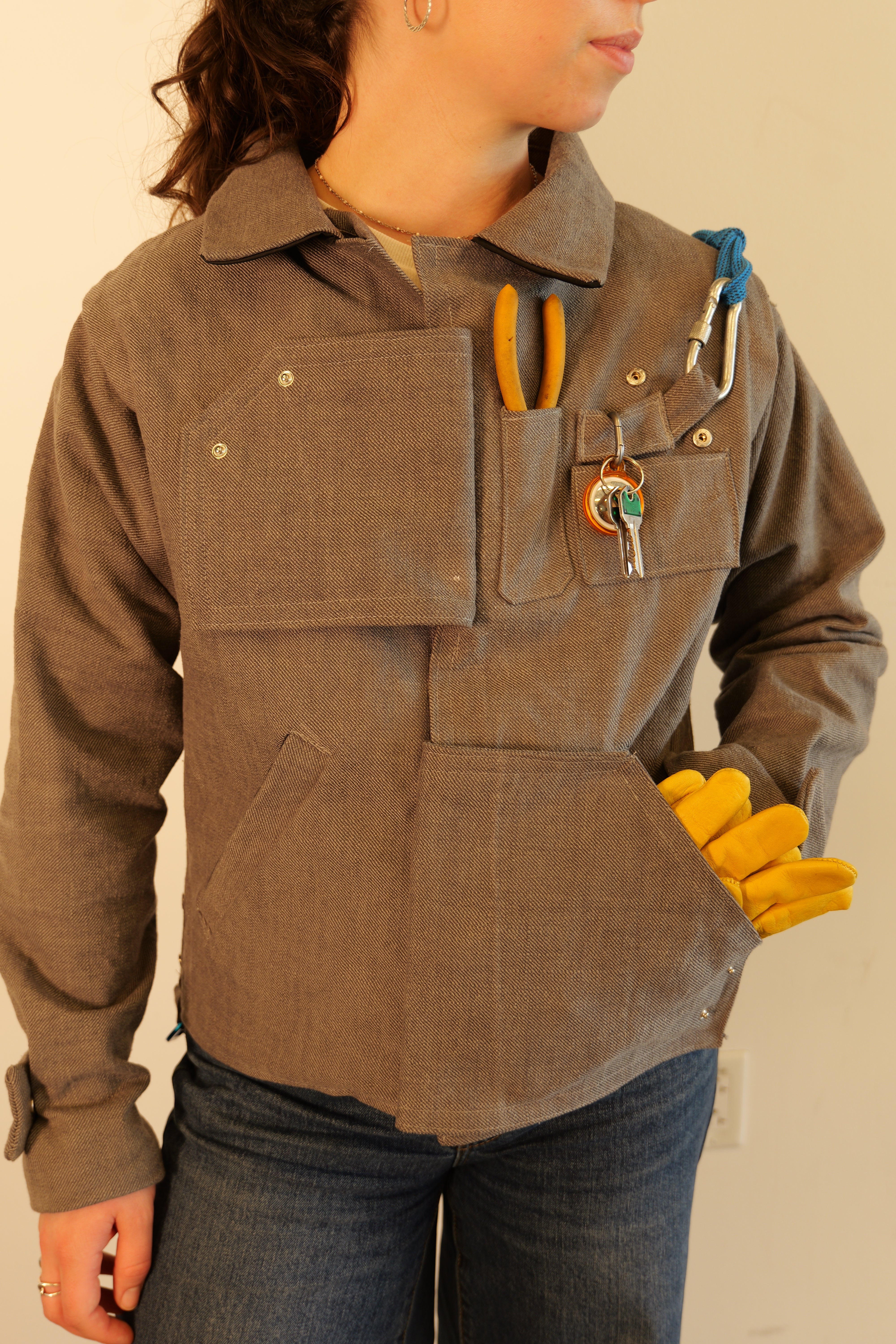 The Open Loop Jacket. A girl wearing a gray, long-sleeve carpenter's jacket with many pockets that hold utilitarian tools. The sleeves of the jacket snap off, and pockets act as the upper and lower closures.