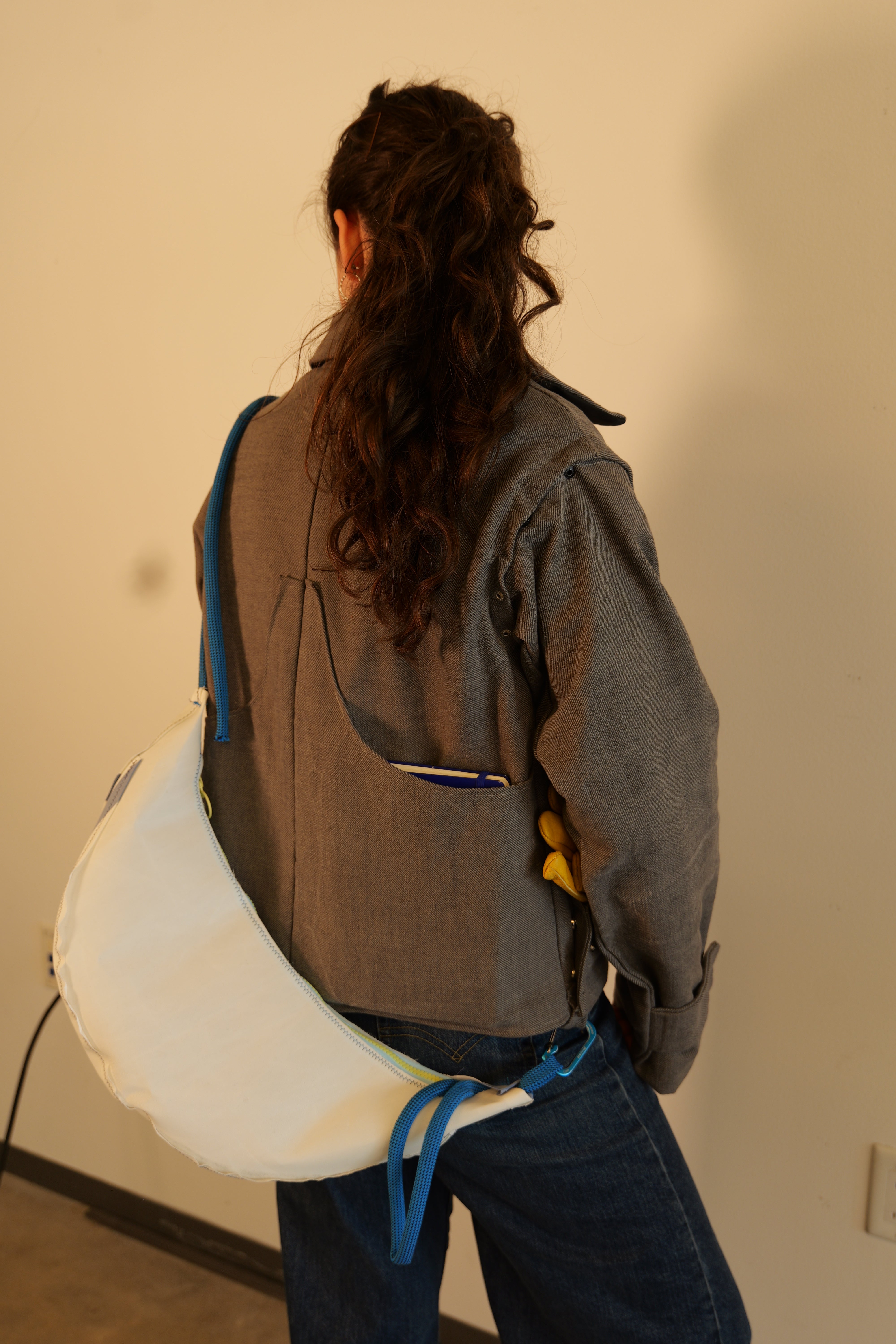 The back of a girl wearing a gray jacket and blue jeans. A white crescent bag with blue rope straps has been hooked into loops on the jacket to sling it across the back. A notebook is placed in the right side panel.