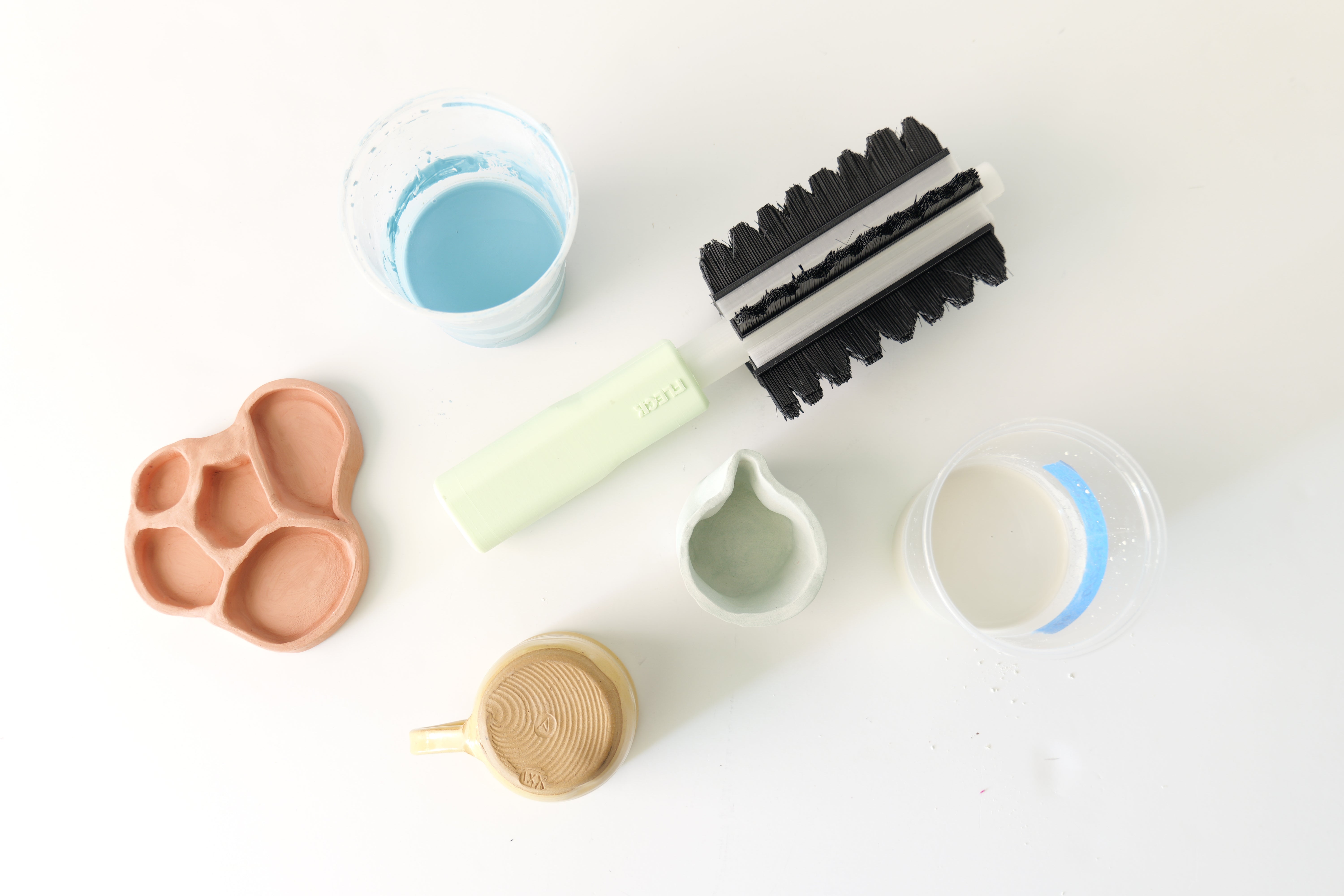 On a white background, a green handled brush with black bristles oriented near several jars of blue, grey, and white ceramic glaze liquid. A yellow mug.