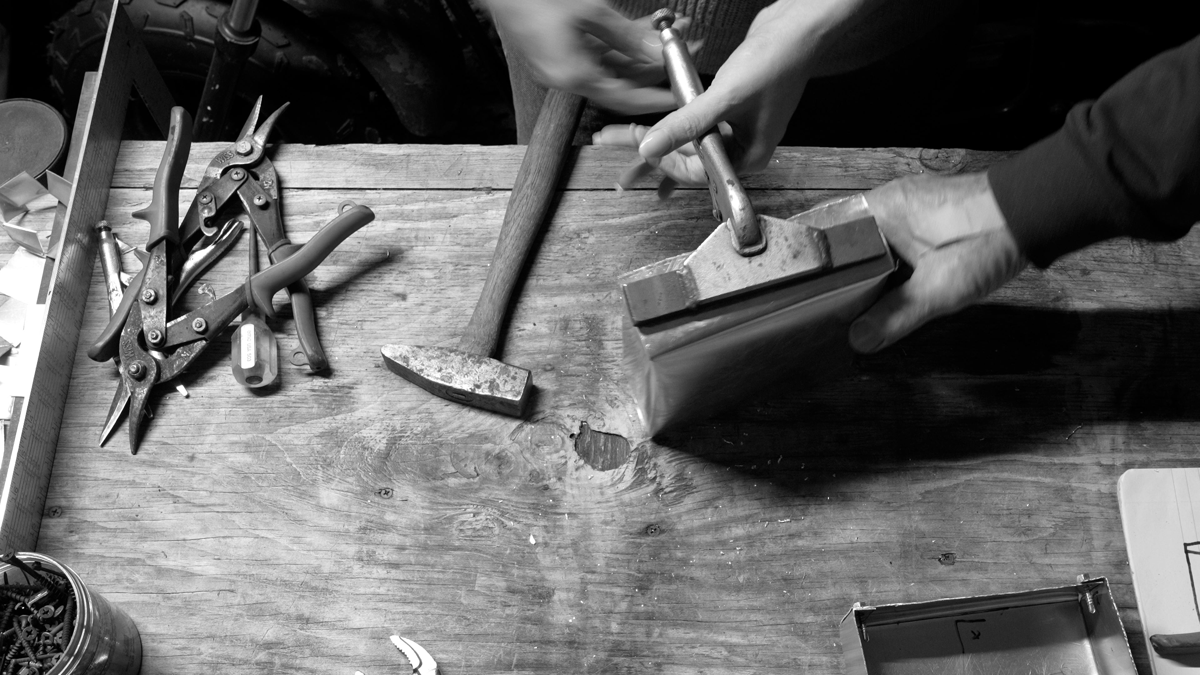 Video still in black and white from a project about the passing of generational knowledge and skill. Two people are shaping a metal box with sheet metal tools.