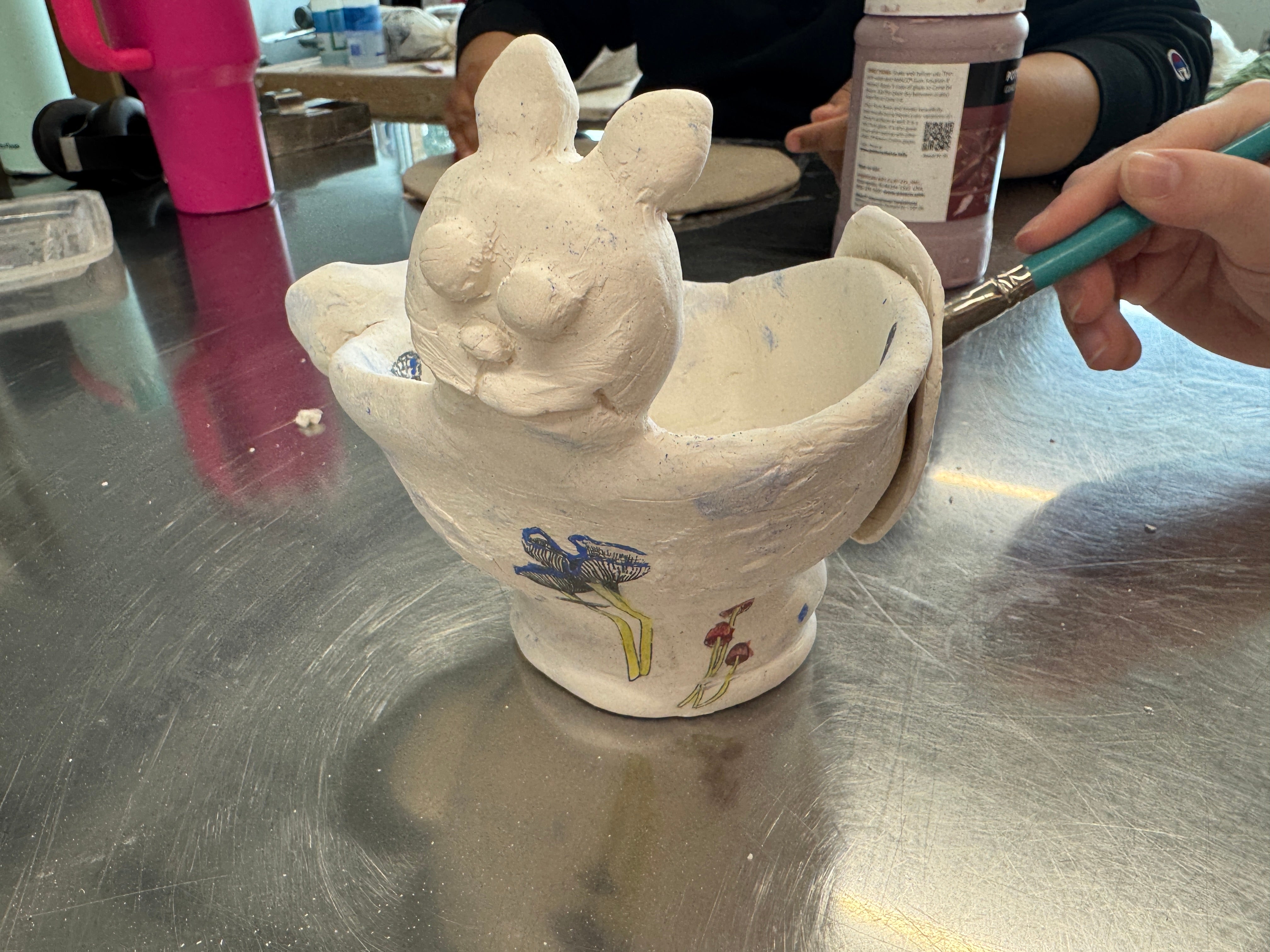 A student out of frame glazes an oversized tea cup with mushrooms and a cat face