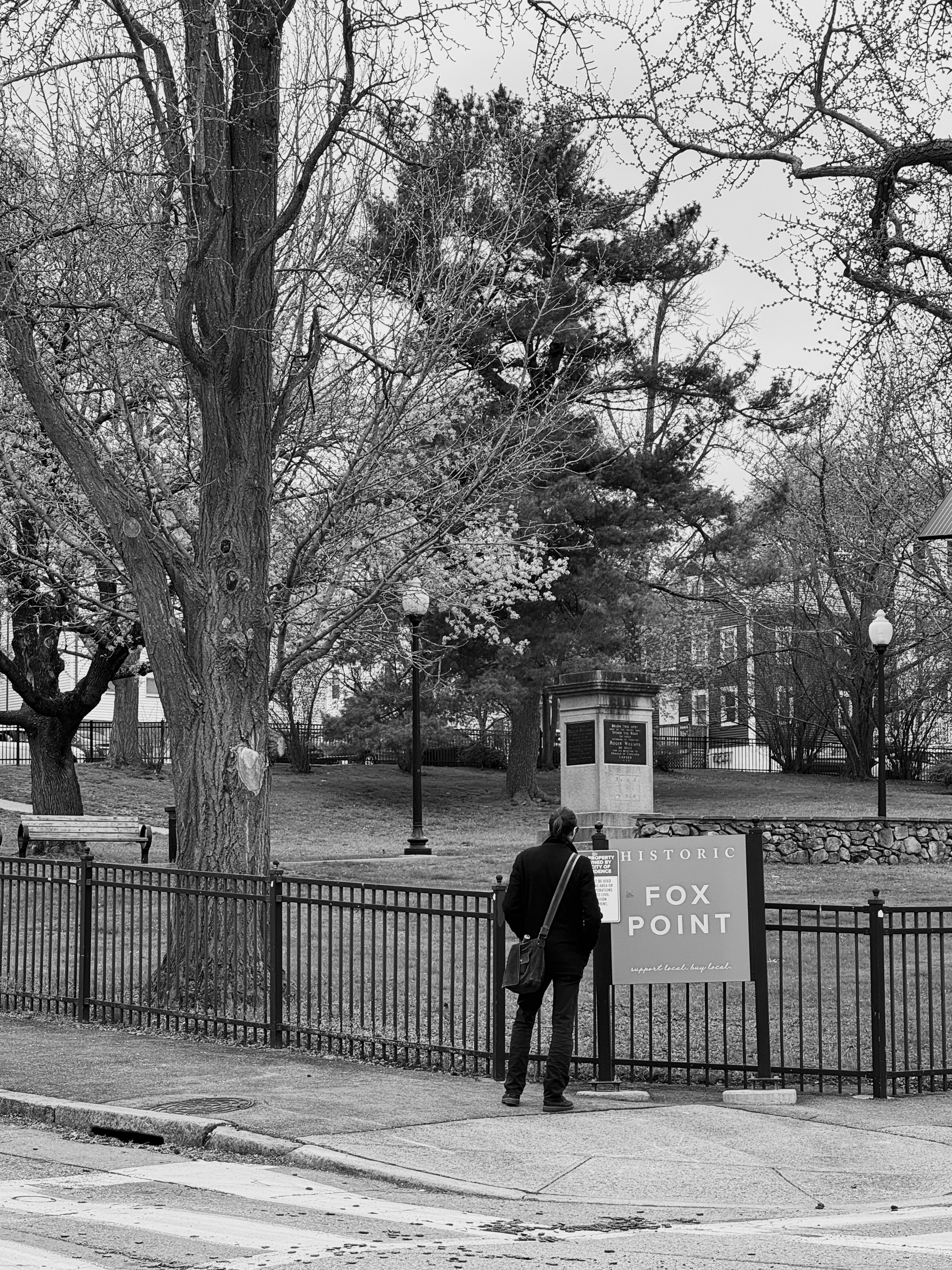 A black-white photo with a man standing back to the camera, looking at a billboard describing an urban green space in the background. He is wearing a black jacket with his hair tied up.