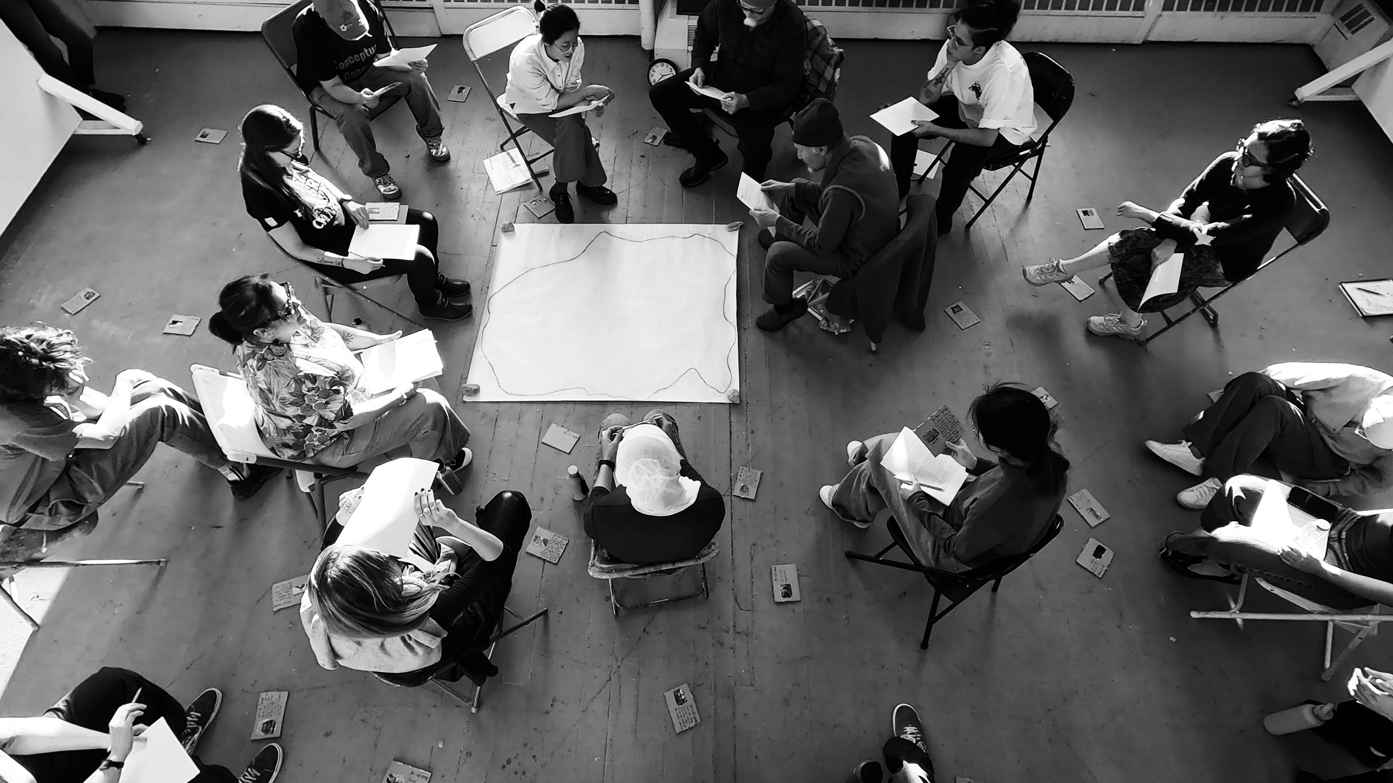 A high-angle black-white shot with 18 people sitting in a circle listening and reading. Three of them are holding printed paper and reading text from it. Paper and cardboards with text and images are scattered around.