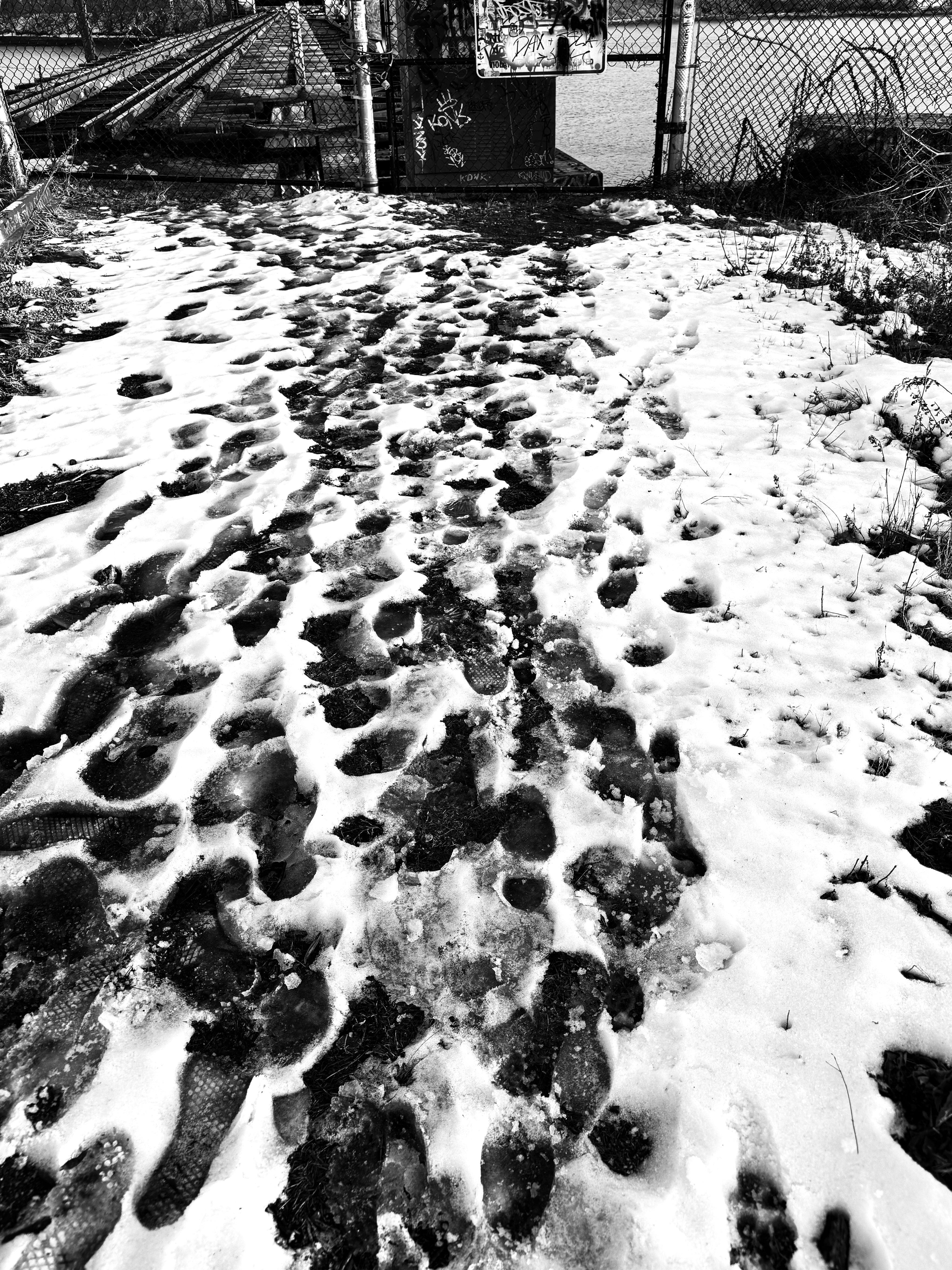 Black-white photo with footprints on the snow pathway directing to a half-open broken iron gate in the distance. Inside the gate, train tracks on a bridge are forwarding.