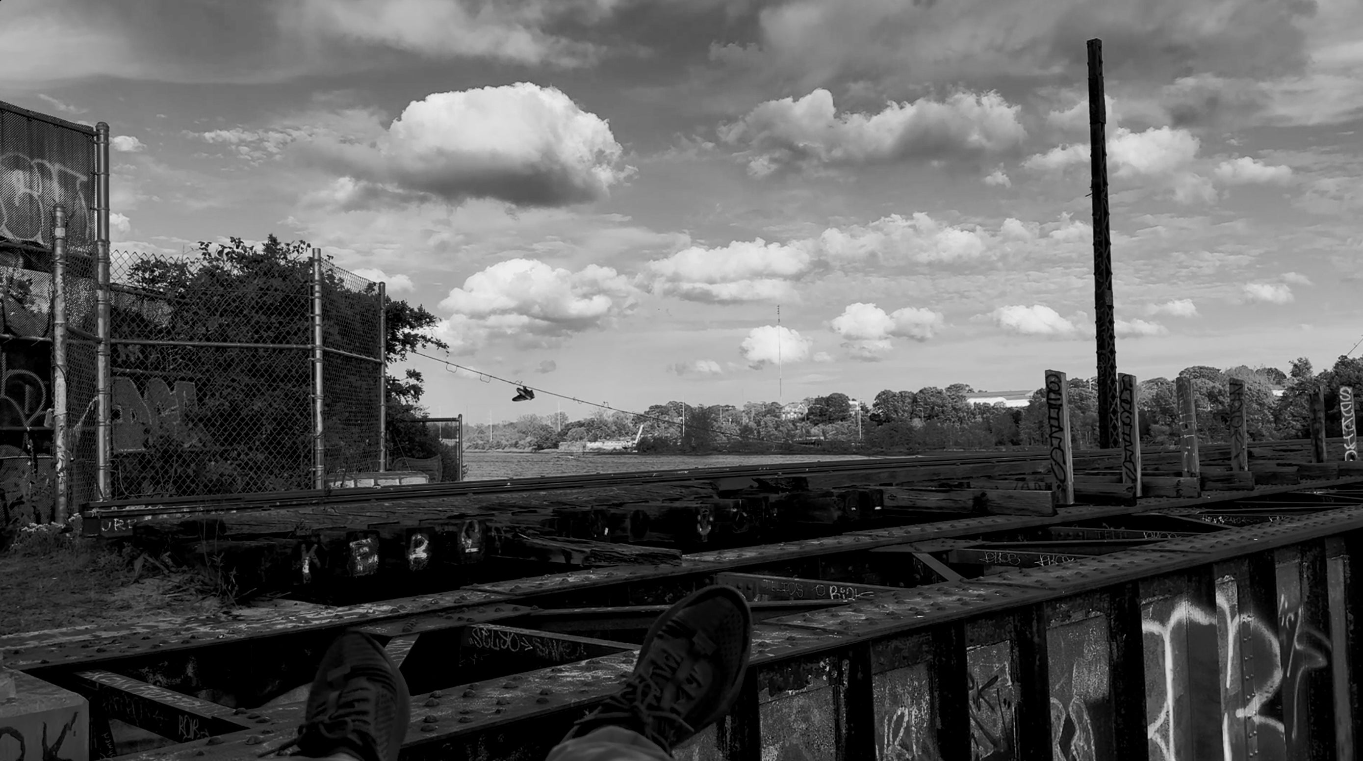 Black-white photo taken from a person’s angle watching over the train track running through the middle of the frame, with feet waving in the foreground. In the distance, a pair of shoes hangs from a rope.