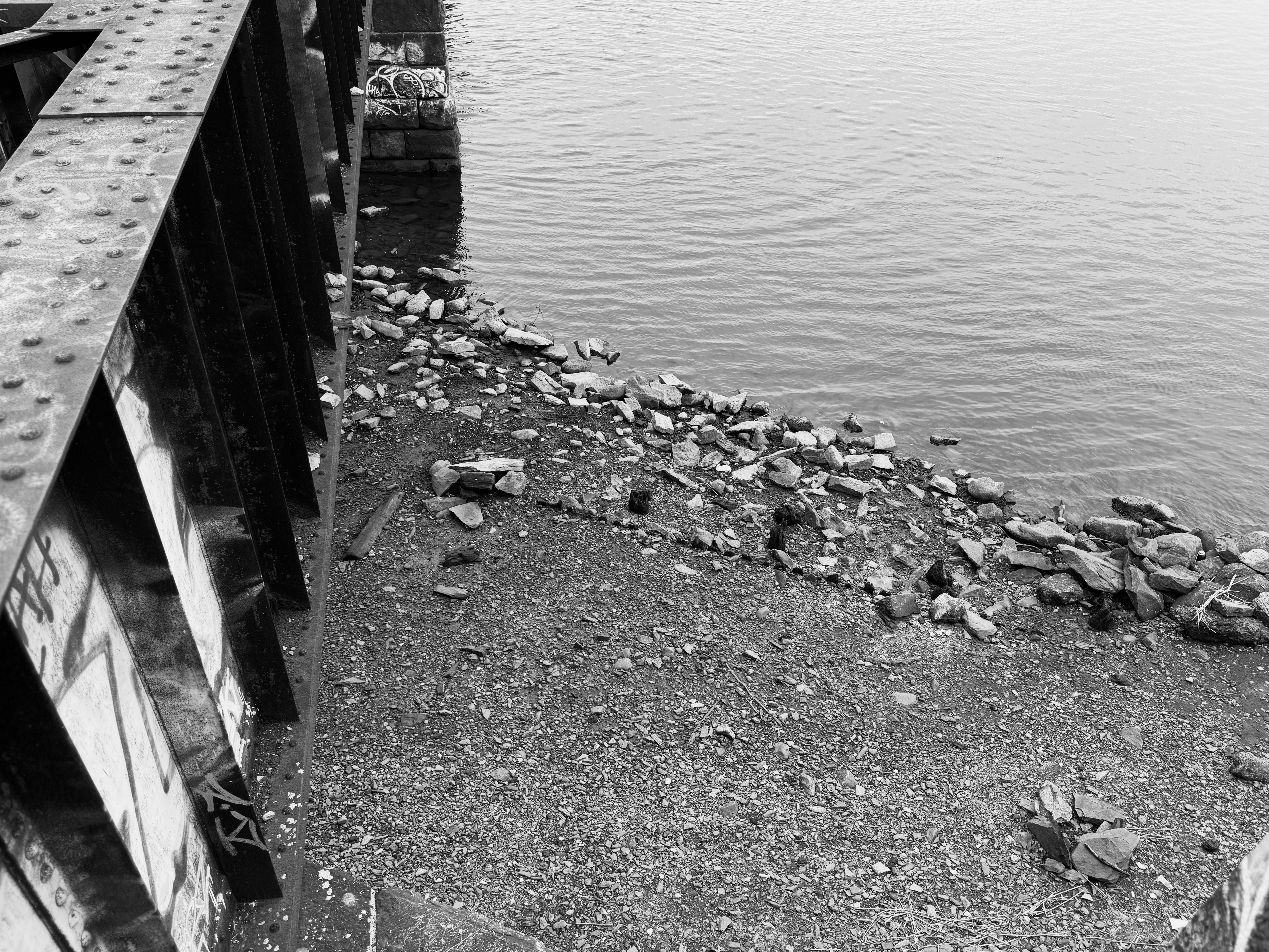 An overhead black-white shot of a rocky shore on the far side of the frame while a part of the train track is leading towards on left front side.