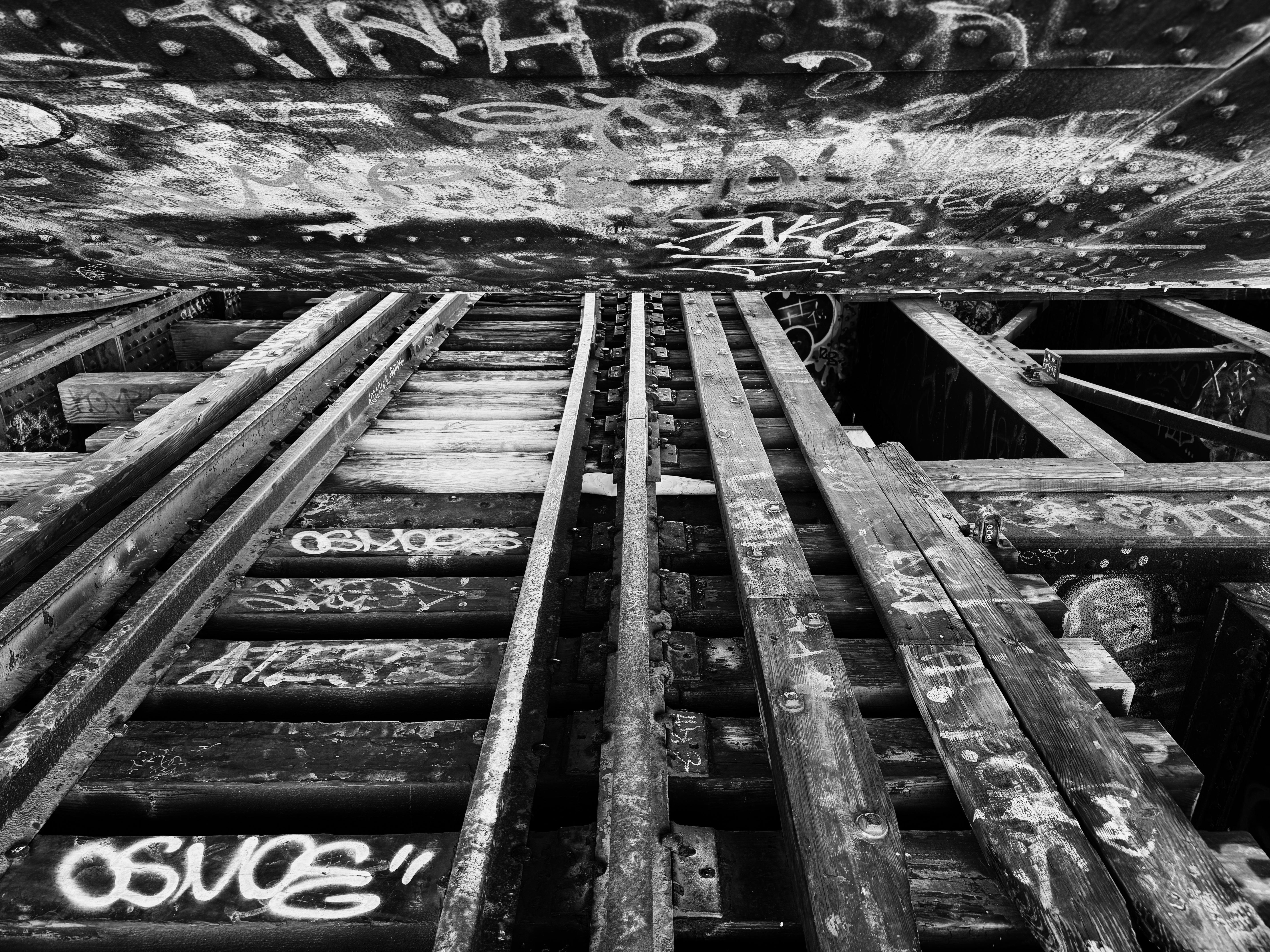 Black-white photo with a stretch of old, abandoned train tracks under an up-structure of a bascule bridge, covered with graffiti and burnt woods. A white sheet of paper is rolled up and embedded in the middle.