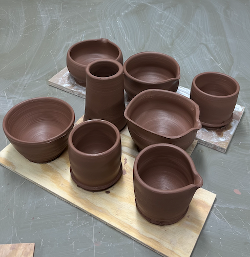 assorted thrown greenware pottery in dark brown clay, resting on dusty table.